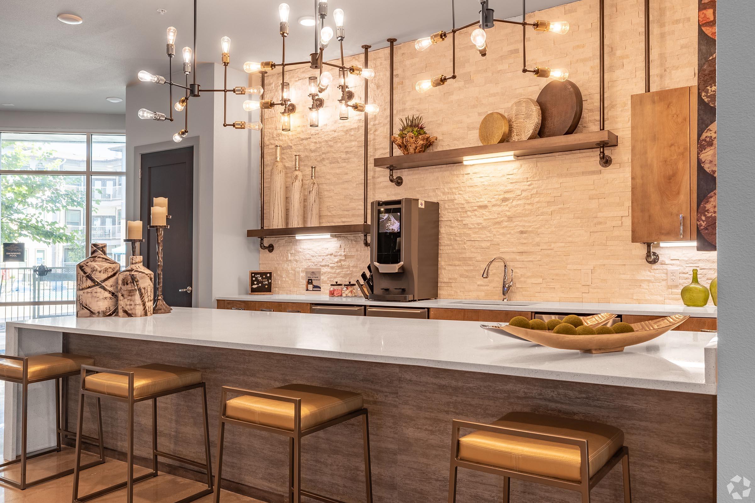 A modern kitchen with a light-colored countertop and wooden cabinetry. There are stylish bar stools along the counter. A unique chandelier hangs from the ceiling, providing warm lighting. The wall features stone accents and decorative elements like bowls and plants, creating an inviting atmosphere.