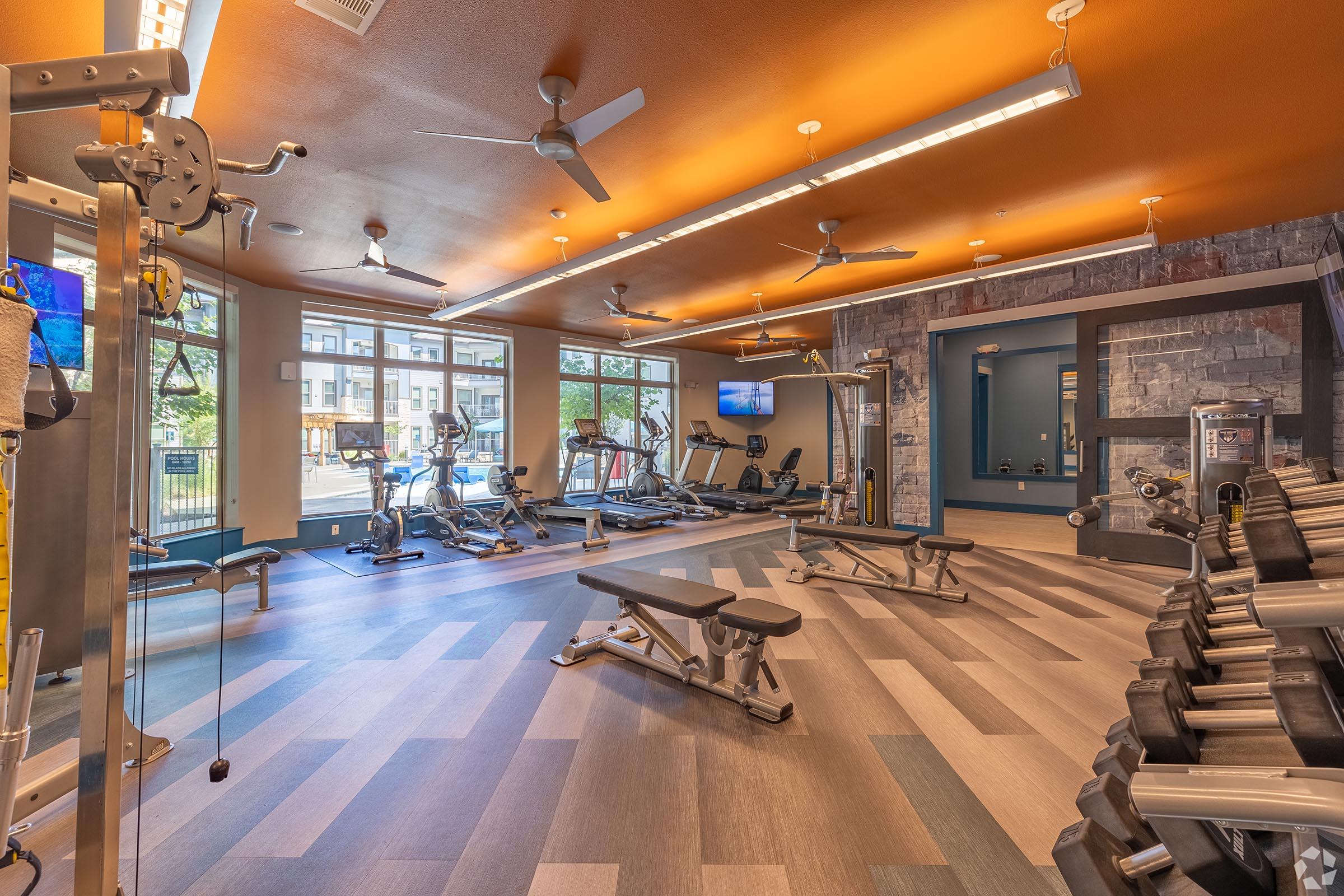 A modern gym interior featuring exercise equipment such as treadmills, weights, and benches. The space has a warm color palette with ceiling fans, large windows allowing natural light, and multiple flat-screen TVs mounted on the walls. The flooring has a contemporary design, enhancing the atmosphere.