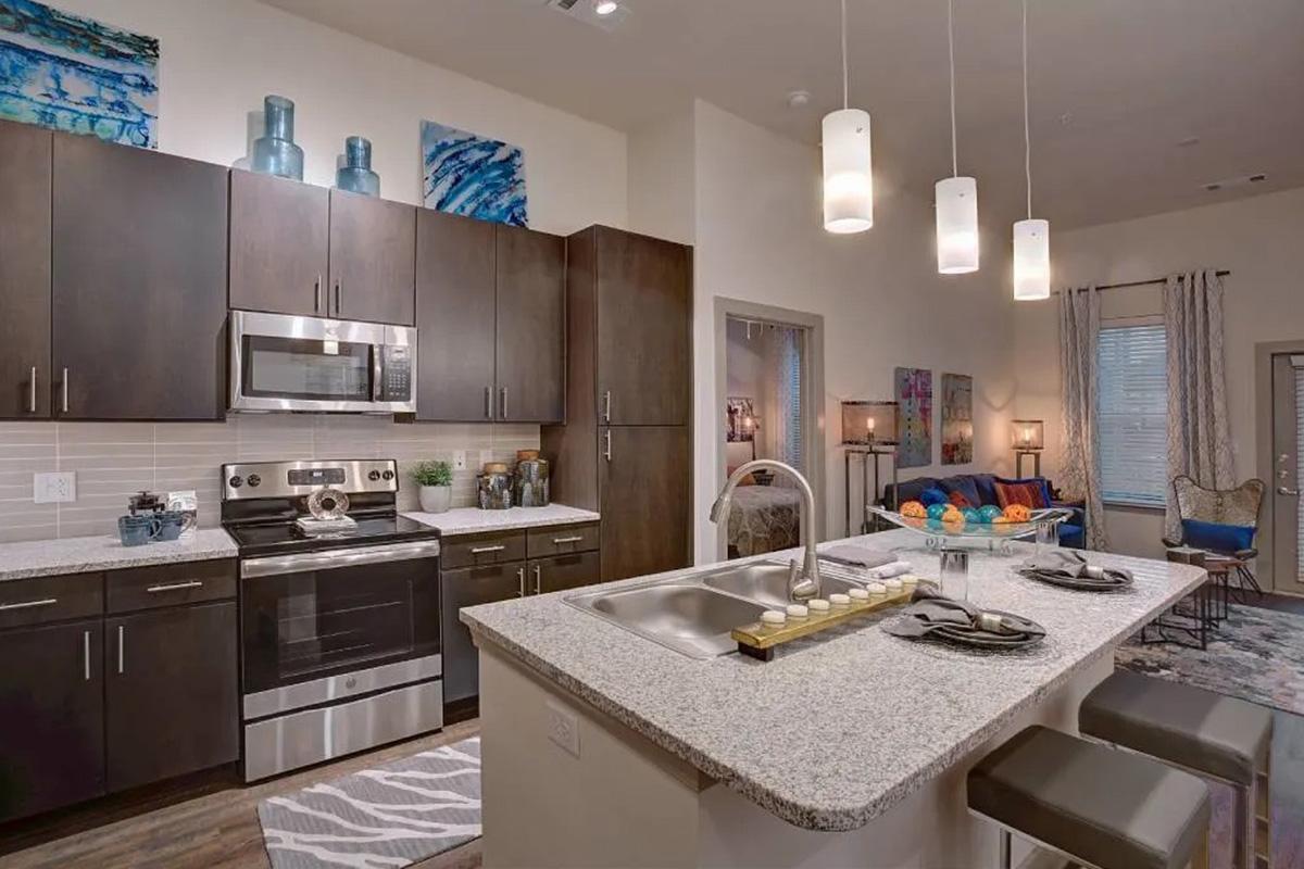Modern kitchen featuring dark wooden cabinets, stainless steel appliances, and a large granite countertop. There are decorative items like blue vases and artwork on the walls. The space is open to a living area with comfortable seating and colorful decor, illuminated by pendant lights.
