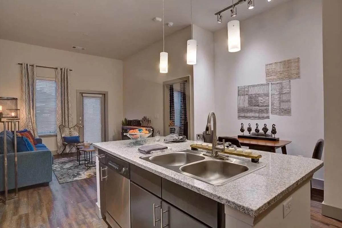 Modern kitchen with a granite island featuring a double sink. Pendant lights hang above, illuminating the space. To the left, a cozy living area with a blue couch and decorative accents. A dining area with a wooden table and decorative items is visible in the background, along with large windows and minimalist wall decor.