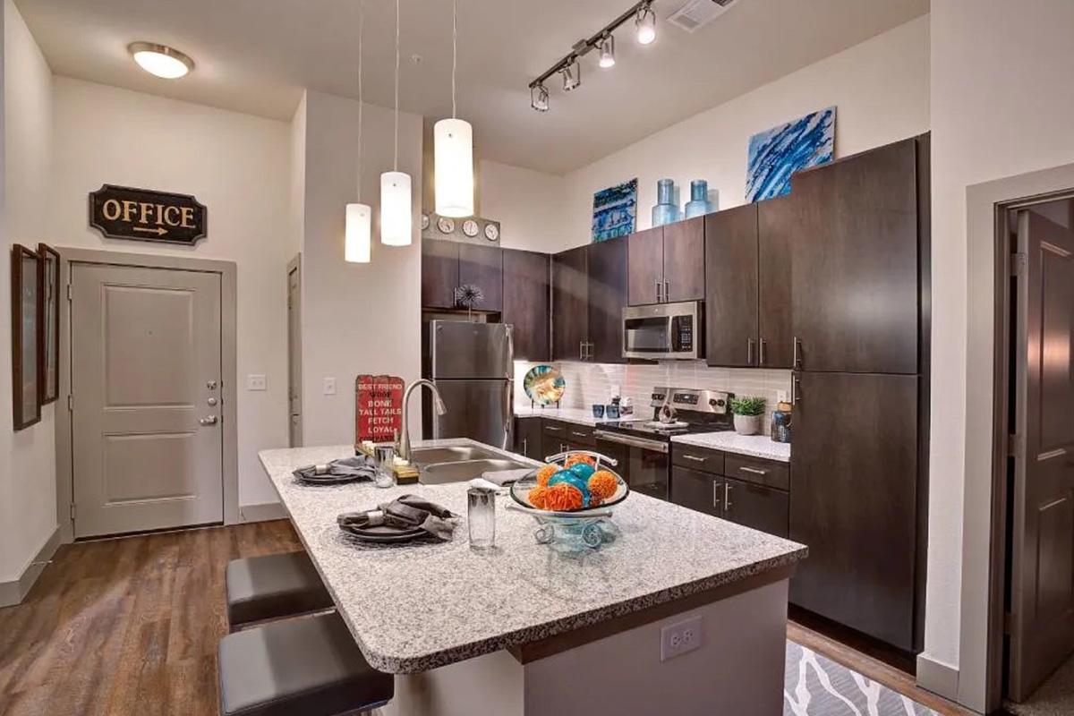 Modern kitchen featuring dark wooden cabinetry and granite countertops, with an island that has bar seating. Decorative accents include colorful bowls and artwork on the walls. Ambient lighting is provided by pendant lights above the island. A doorway leads to an office, visible in the background.