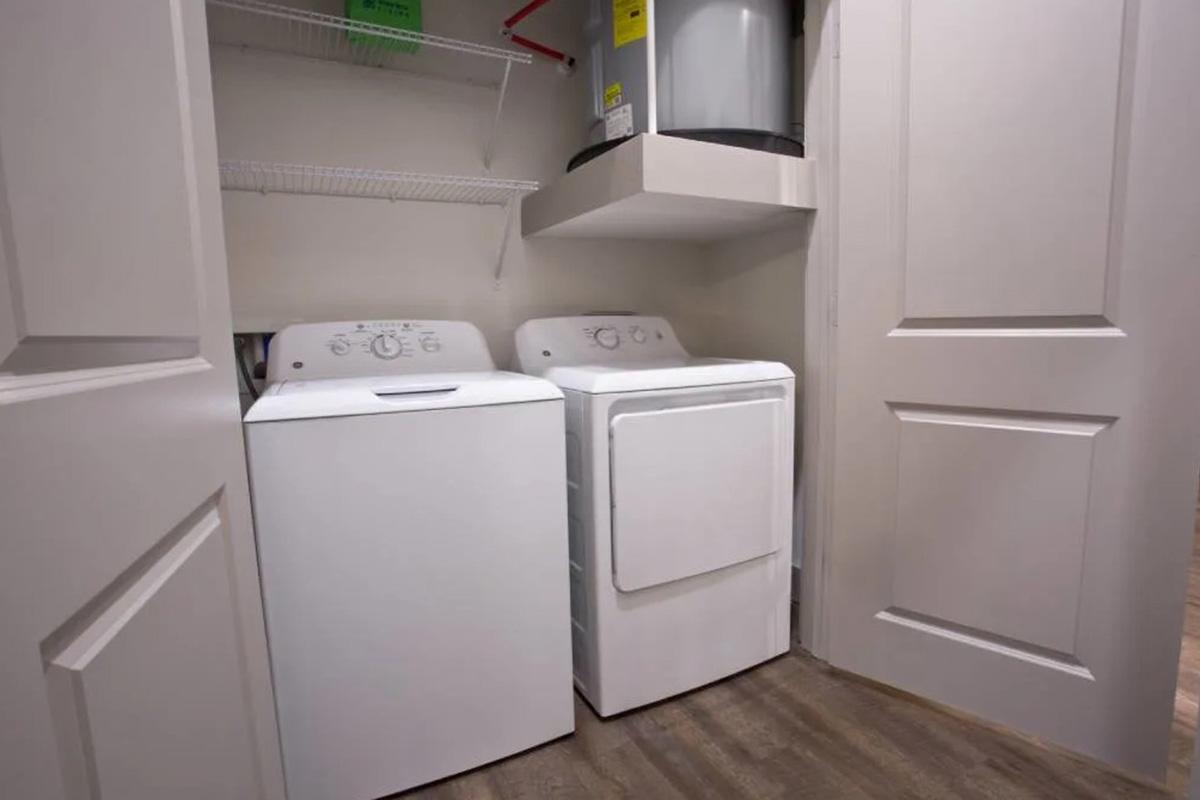 A laundry room featuring a white washer and dryer side by side, positioned within a small space. There are shelves above the appliances and a water heater or other appliance visible in the corner. The room has beige walls and a wooden floor. The doors to the room are partially closed.