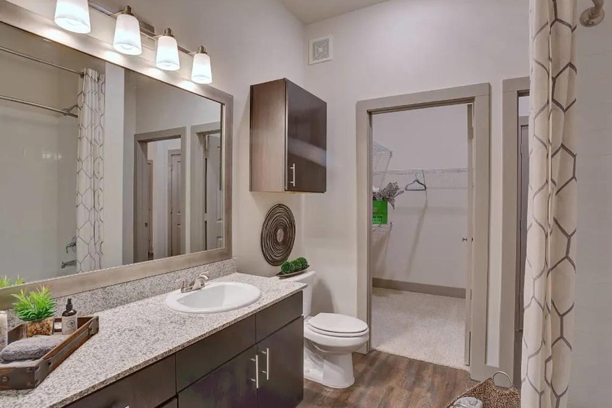 A modern bathroom featuring a granite countertop with a sink, a large mirror, and dark wood cabinetry. The space includes a toilet, textured flooring, and minimal decor. A door leads to a closet area with a wire rack. Soft lighting from above complements the neutral color scheme.