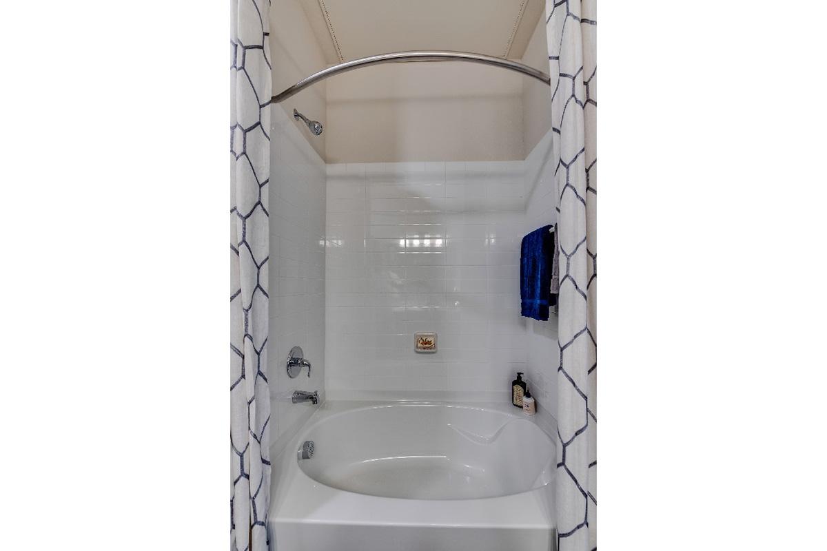 A modern bathroom featuring a curved shower curtain rod enclosing a bathtub with white tile walls. A blue washcloth hangs on the side, and a small bottle of shampoo or body wash is placed near the faucet. The overall design is clean and simple, with a bright and airy feel.