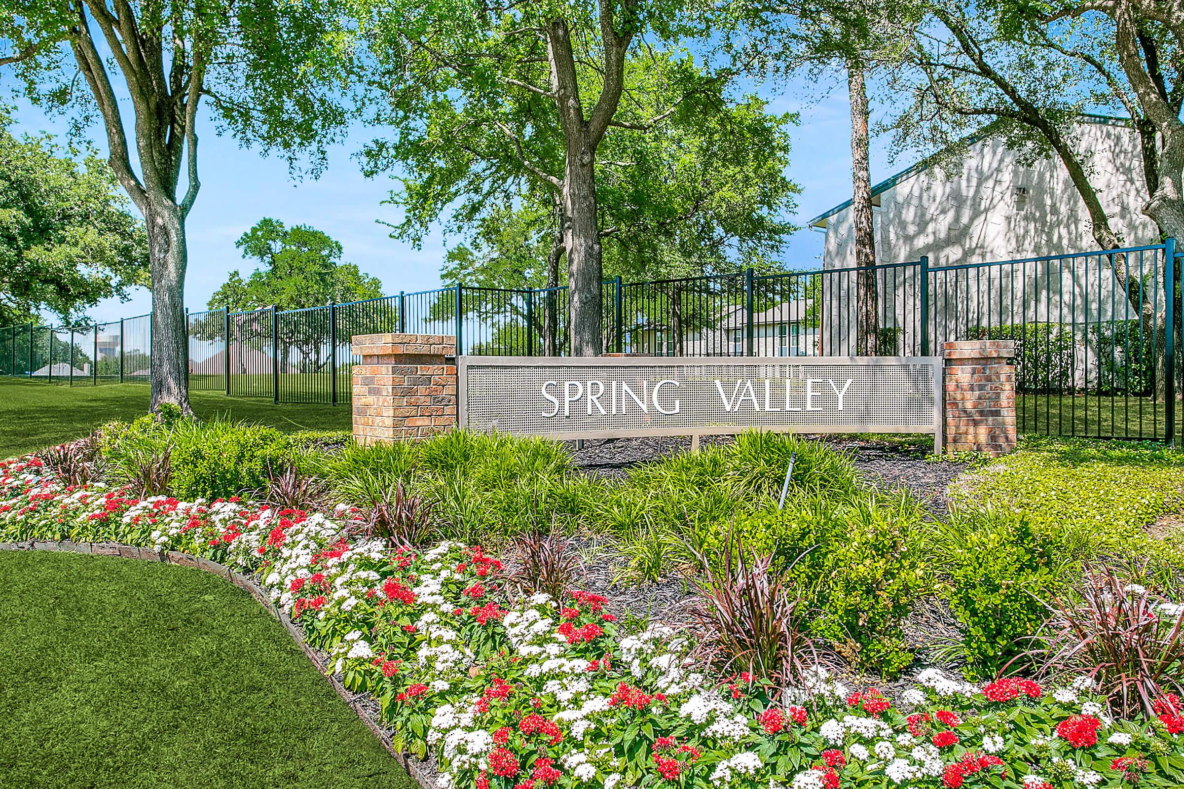 A neatly landscaped entrance sign for "Spring Valley," featuring a stone and brick structure with white lettering. The sign is surrounded by vibrant flower beds in various colors, with green grass and trees in the background, creating a welcoming atmosphere.