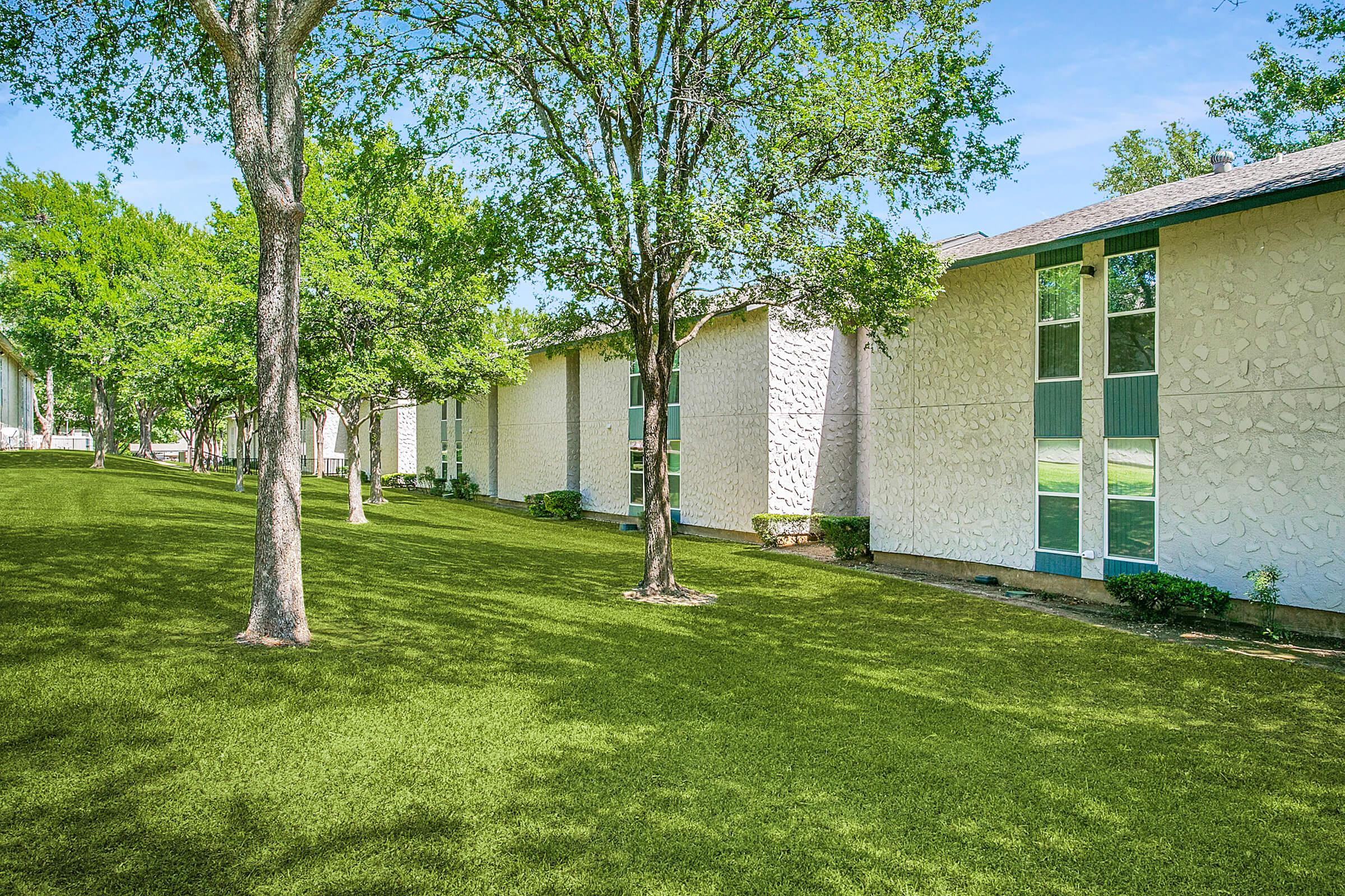 A landscaped area featuring a row of modern buildings with textured walls, surrounded by lush green grass and trees. The scene is bright and sunny, showcasing a well-maintained outdoor space suitable for leisure or recreation.