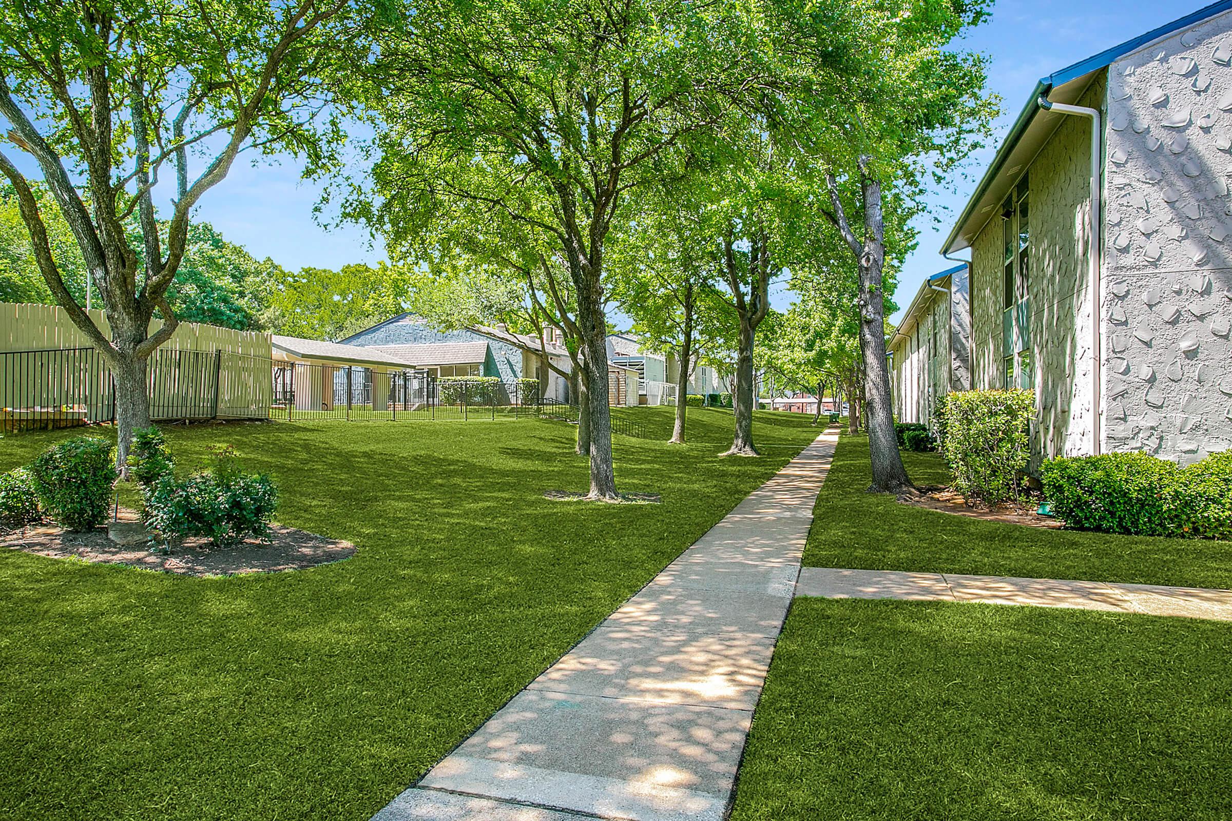 A well-maintained outdoor area featuring a paved walkway lined with trees and greenery. Surrounding buildings are visible in the background, with well-trimmed grass and a clear blue sky above, creating a pleasant and inviting atmosphere.