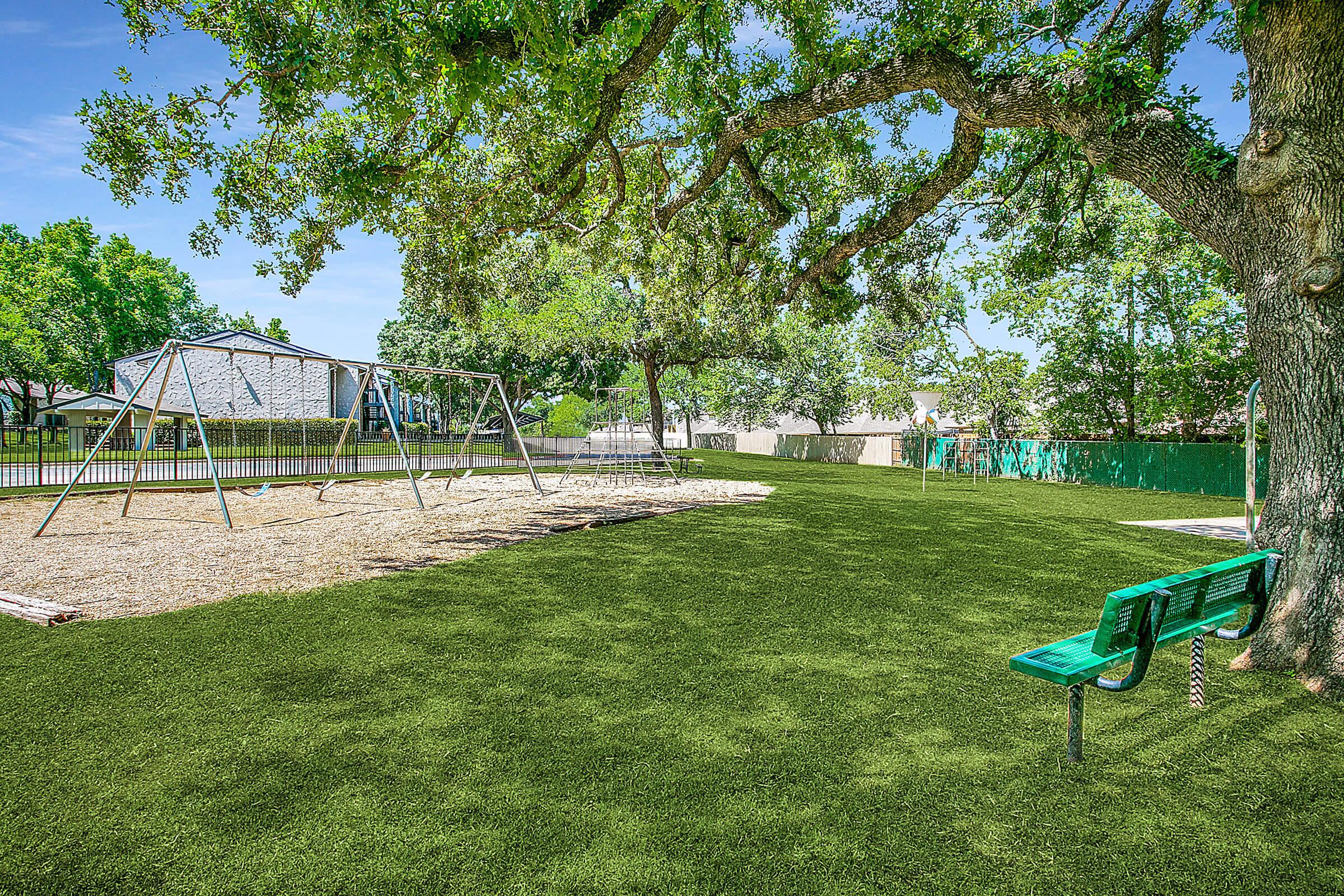 A playground featuring swings and a green bench in a park. The ground is covered in artificial grass, and there are lush trees providing shade. The background includes a fenced area with additional greenery, creating a peaceful outdoor space for recreation and relaxation.