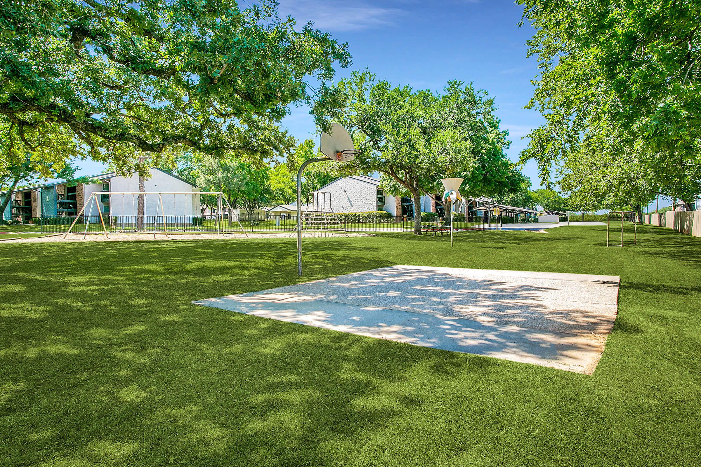 A sunny park scene featuring a grassy area with two basketball hoops, a playground with swings, and shaded trees. Bright blue sky and distant buildings are visible, creating a cheerful, inviting atmosphere for outdoor activities and enjoyment.