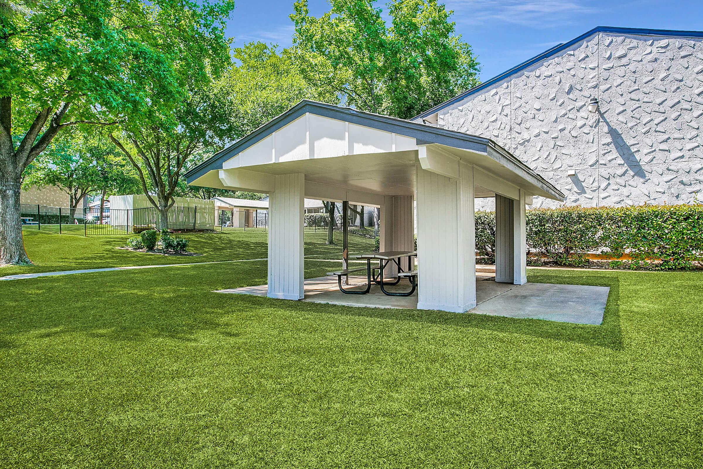 A covered picnic area with a table is situated on lush green grass, surrounded by trees. In the background, a light-colored building and additional greenery are visible, creating a relaxing outdoor space.