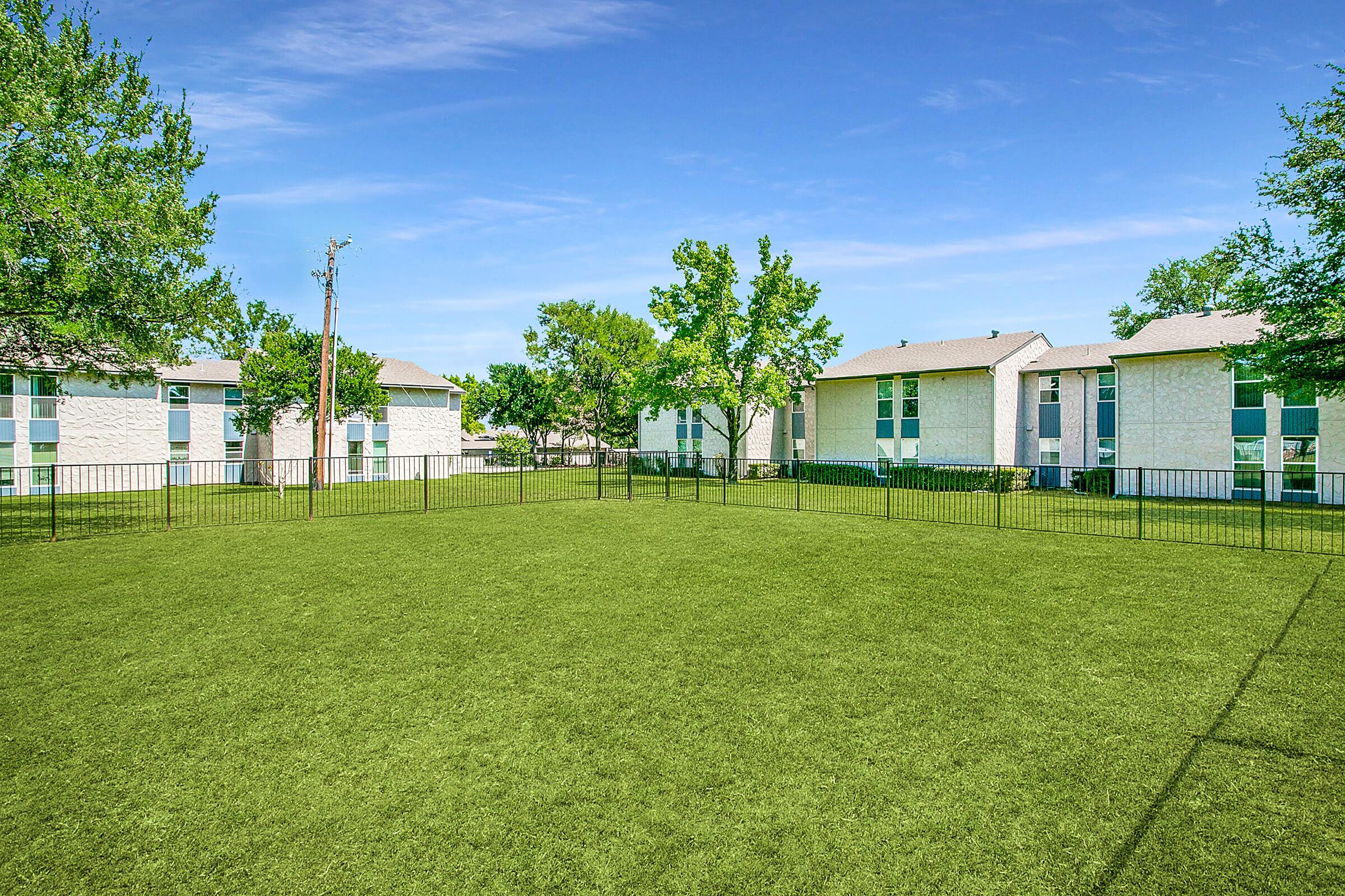 A large, green outdoor space surrounded by low black fencing, featuring trees and residential buildings in the background under a clear blue sky. The area appears well-maintained and is suitable for recreational activities.