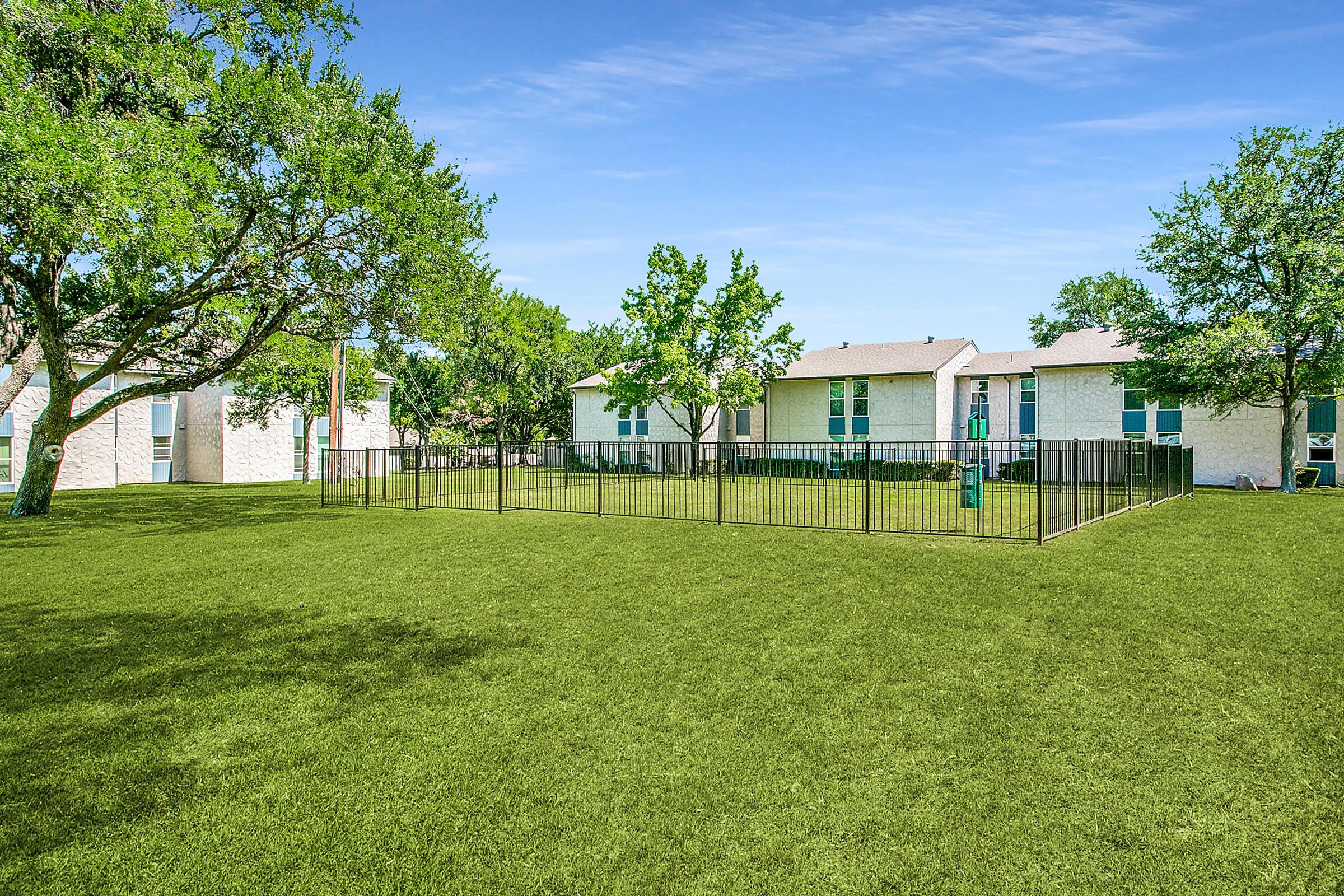 A spacious green lawn enclosed by a black fence, surrounded by trees. In the background, there are simple, light-colored buildings with multiple windows. The sky is clear and blue, creating a bright, inviting atmosphere.