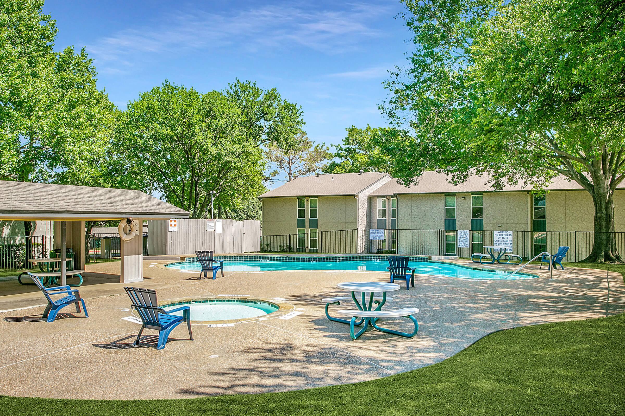A sunny outdoor pool area featuring a sparkling blue pool and a hot tub, surrounded by green trees. There are several blue lounge chairs and tables arranged around the pool, with a nearby building in the background. The scene is bright and inviting, perfect for relaxation and leisure.