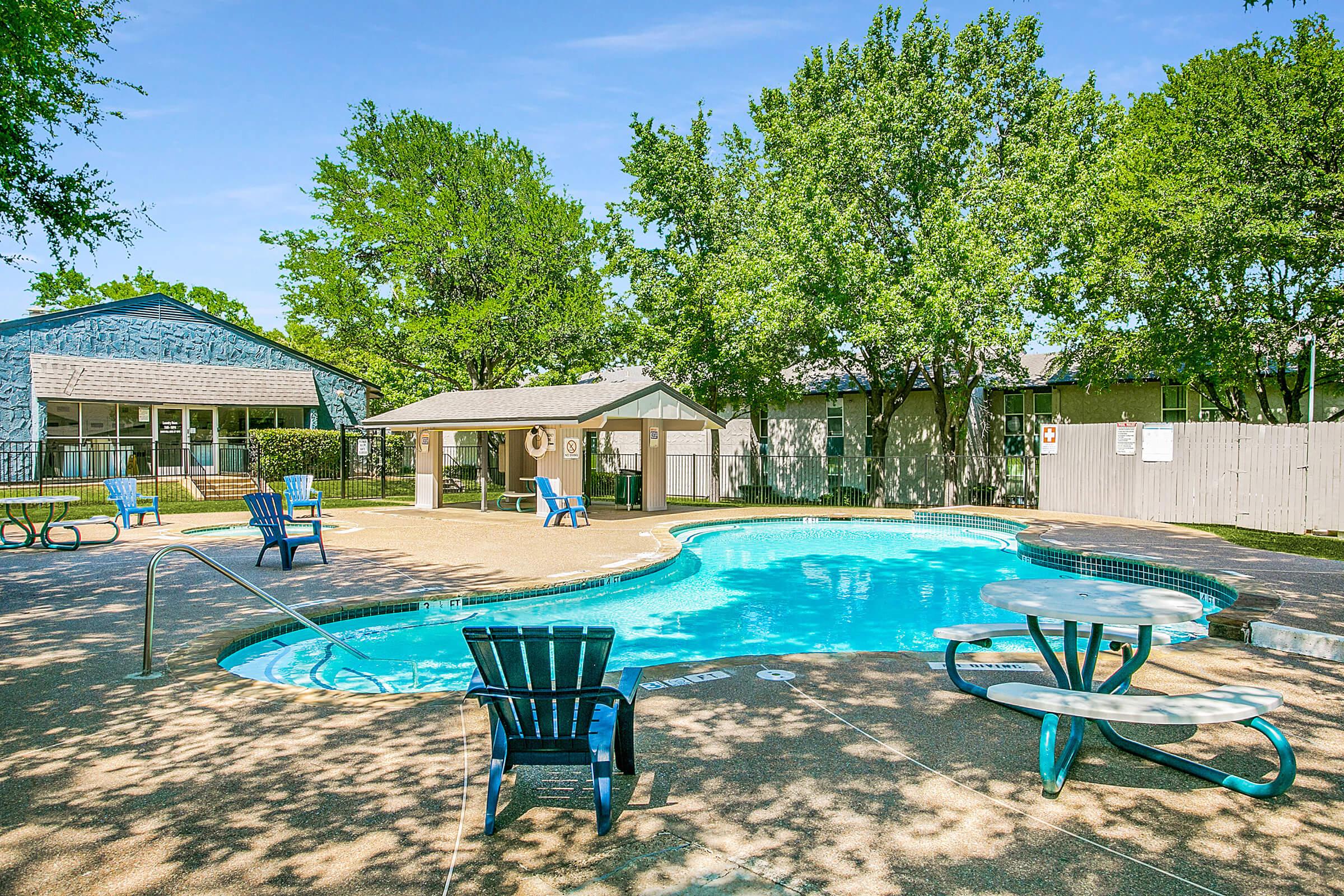 A serene outdoor pool area surrounded by green trees. The pool features a smooth, curvy shape with lounge chairs nearby. There are picnic tables for seating, a shaded gazebo, and a light-colored building in the background. The scene is bright and inviting, perfect for relaxation and leisure activities.