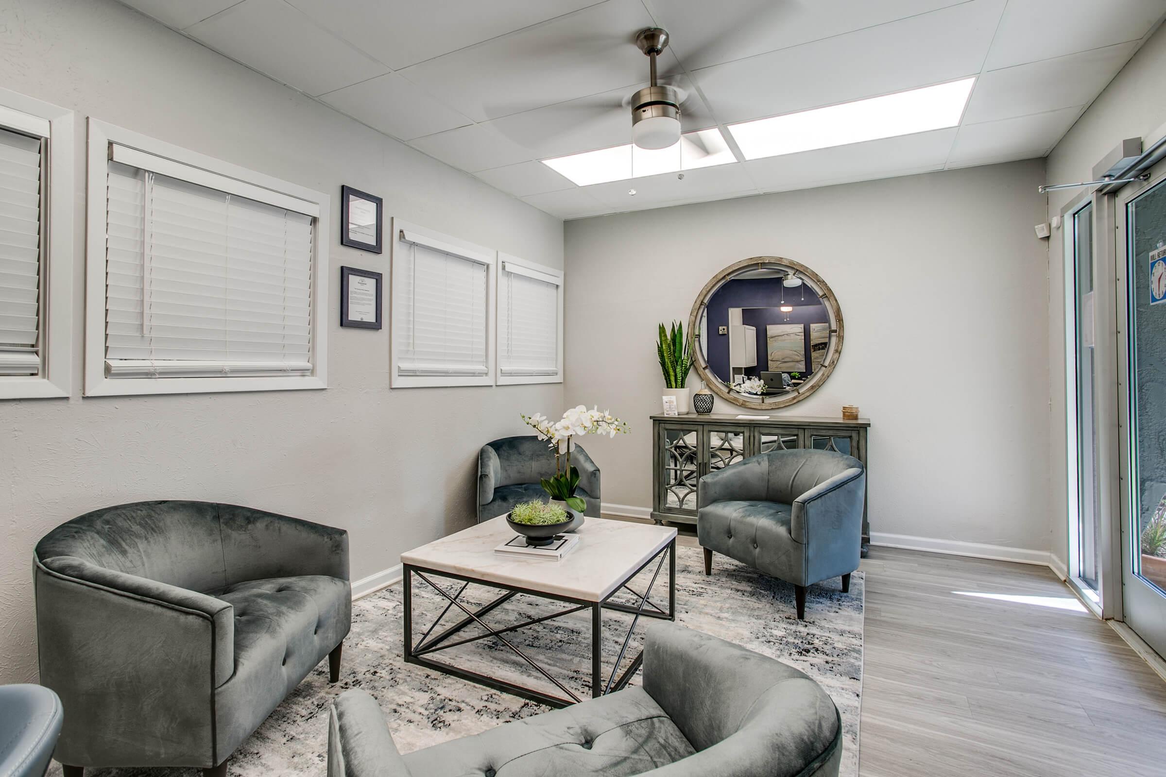 A modern waiting room featuring a light gray color scheme. There are four plush chairs arranged around a coffee table with a decorative plant and a bowl. A round mirror hangs on the wall, and there are large windows allowing natural light to enter. The space has a clean, inviting atmosphere.