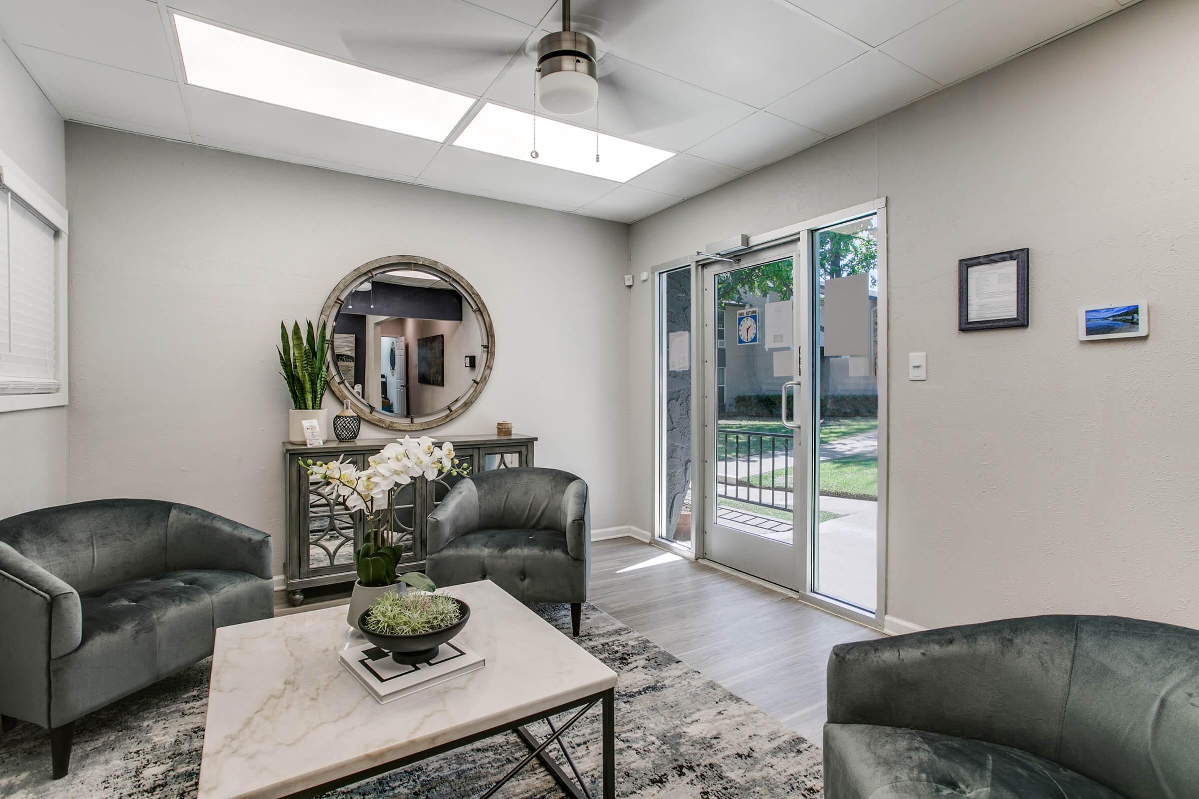 A modern living room with two plush gray chairs, a round mirror, and a decorative coffee table with a plant. Natural light streams in through a skylight and the glass entrance door, leading to an outdoor area. Greenery is visible outside, enhancing the inviting atmosphere.