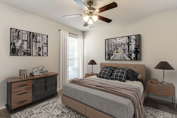 A modern bedroom featuring a queen-sized bed with gray and black bedding, a stylish nightstand on each side with lamps, and a wooden dresser. Two framed artworks hang on the walls. The room has a ceiling fan and a large window covered with sheer curtains, allowing natural light to filter in.