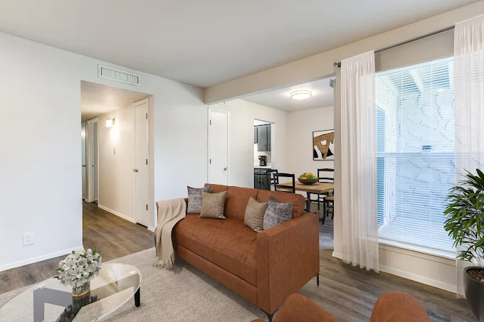 A modern living room featuring a brown sofa with decorative pillows, a glass coffee table, and a light-colored area rug. Natural light streams through large windows with sheer curtains. In the background, a dining area with a table and chairs is visible, along with a doorway leading to a kitchen.