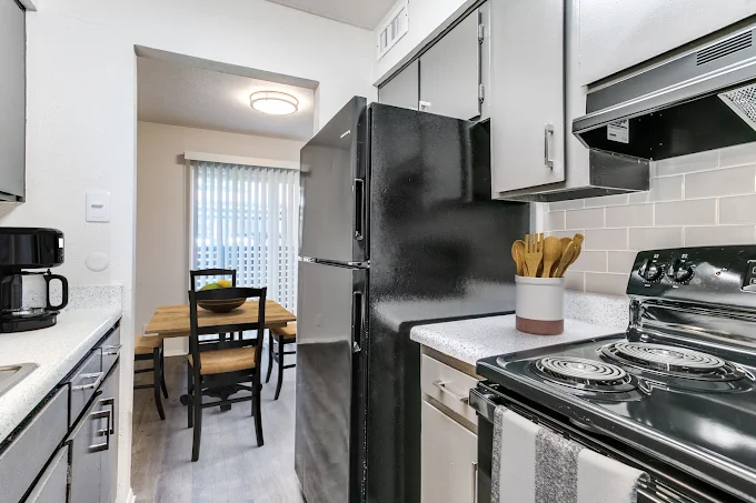 A modern kitchen featuring a black refrigerator, a silver stove with an oven, and a coffee maker on the counter. The kitchen has gray cabinets and a light-colored backsplash. In the background, there is a dining area with a wooden table and black chairs, illuminated by natural light from a window with blinds.