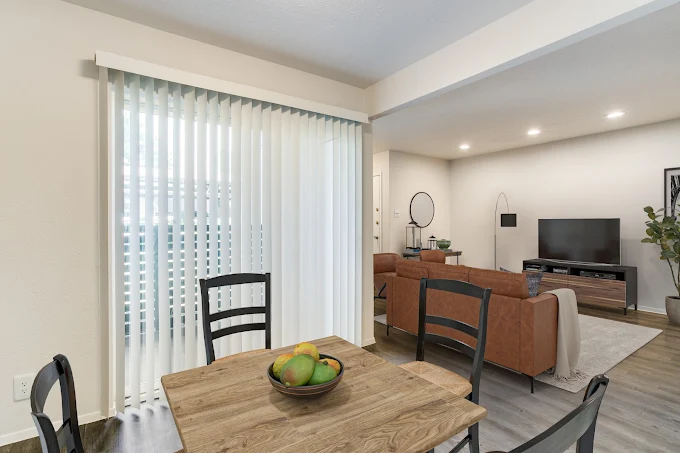A cozy living area featuring a wooden dining table with a bowl of fruit, black chairs, and a living room with a brown couch, TV stand, and decorative plant. There are vertical blinds covering a window, and the space is well-lit with modern lighting, creating a warm and inviting atmosphere.