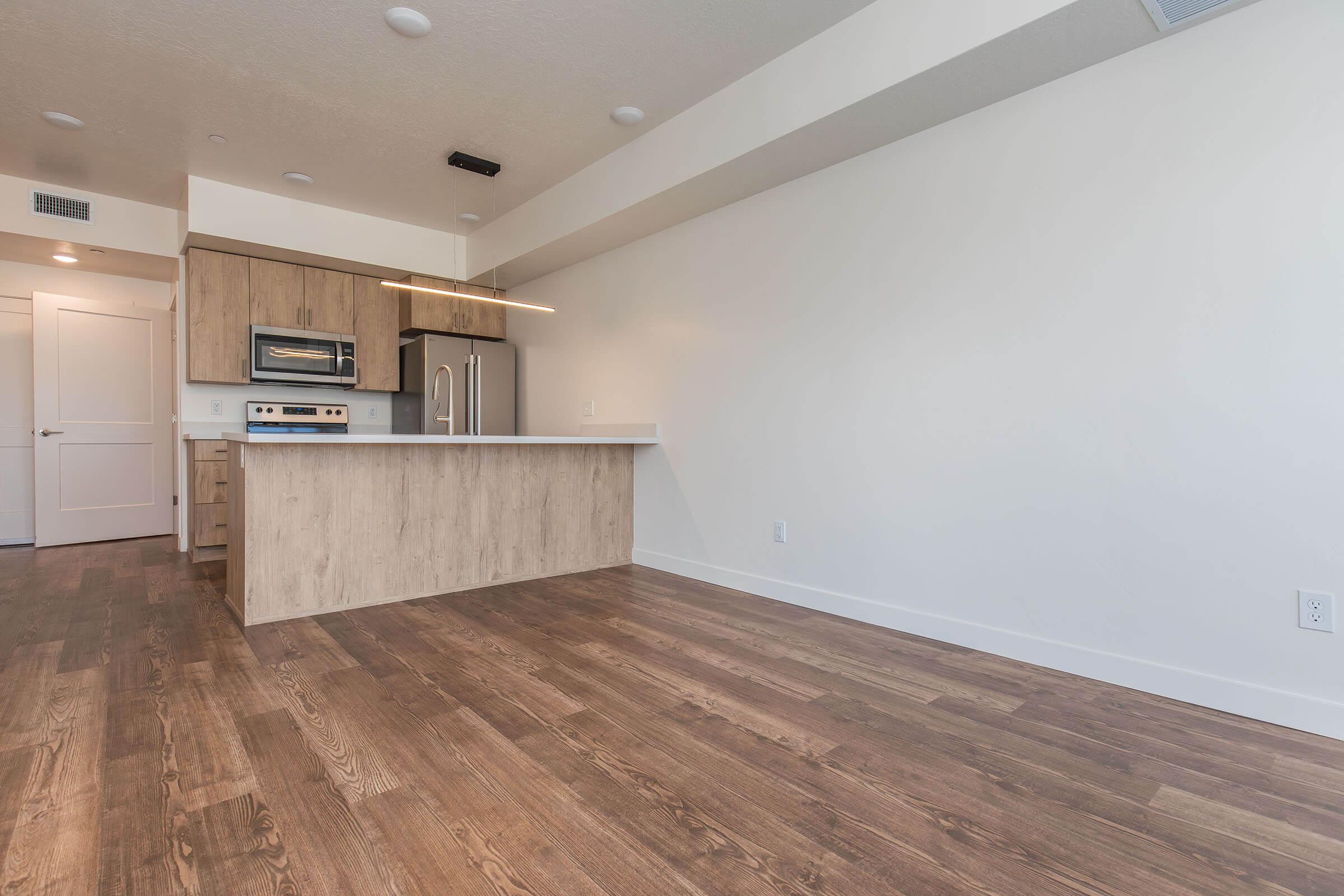 Modern kitchen and living area featuring light wood cabinets, stainless steel appliances, and a white countertop. The room has a light-colored wall and wooden flooring. An island separates the kitchen from the living space, with natural light streaming in through unseen windows.