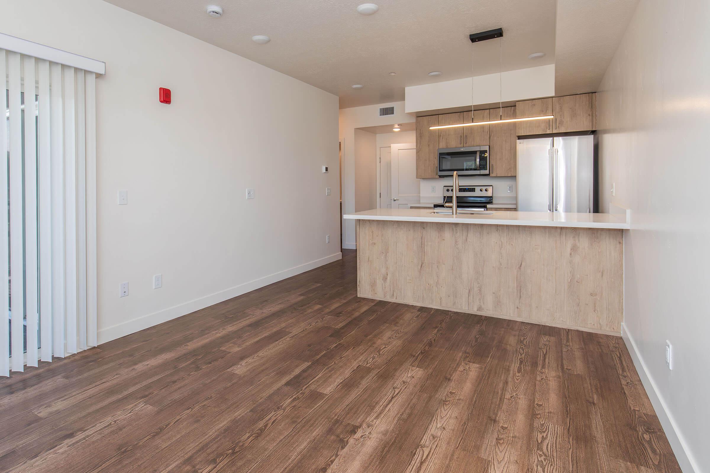 Modern kitchen and living area featuring a light wood floor, white walls, and minimalist design. The kitchen includes a large island with a countertop, stainless steel appliances, and wooden cabinetry. A sliding glass door with vertical blinds leads to outdoors. Bright and spacious with overhead lighting.