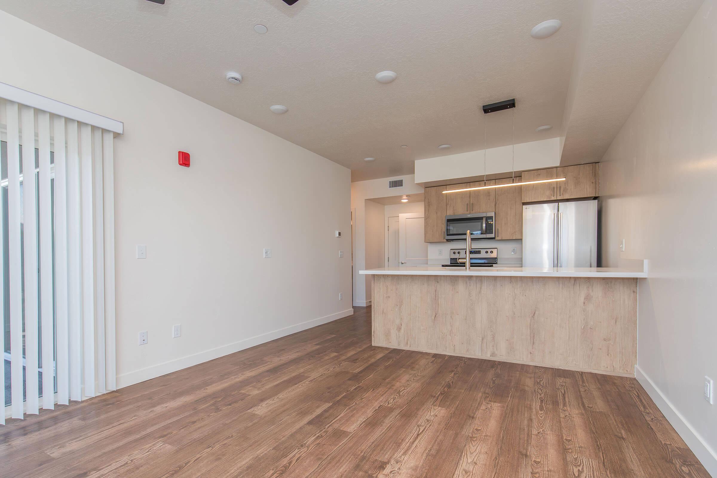 Modern interior of an apartment showing a kitchen with stainless steel appliances, a large island counter, and wooden cabinetry. The living area features wooden flooring and large windows with blinds. The color scheme is light and neutral, creating a spacious and welcoming atmosphere.