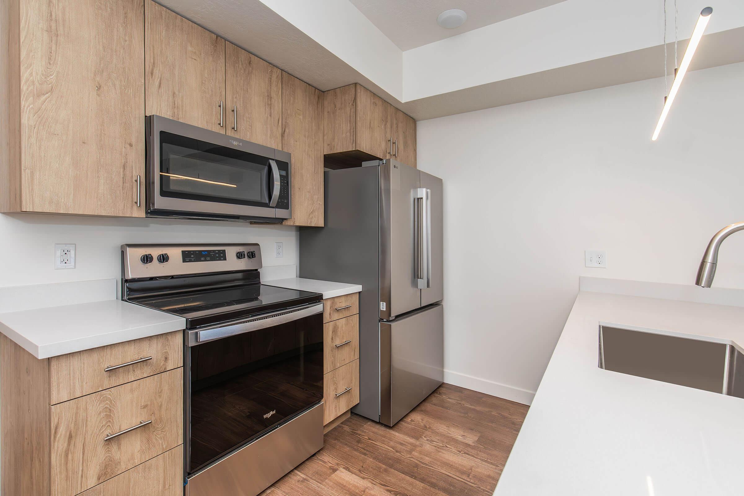Modern kitchen featuring stainless steel appliances, including a microwave, oven, and refrigerator. The cabinetry is light wood, and the countertops are white with a smooth finish. A sleek undermount sink is visible, and the flooring is a light brown wood. The space is well-lit and contemporary in design.