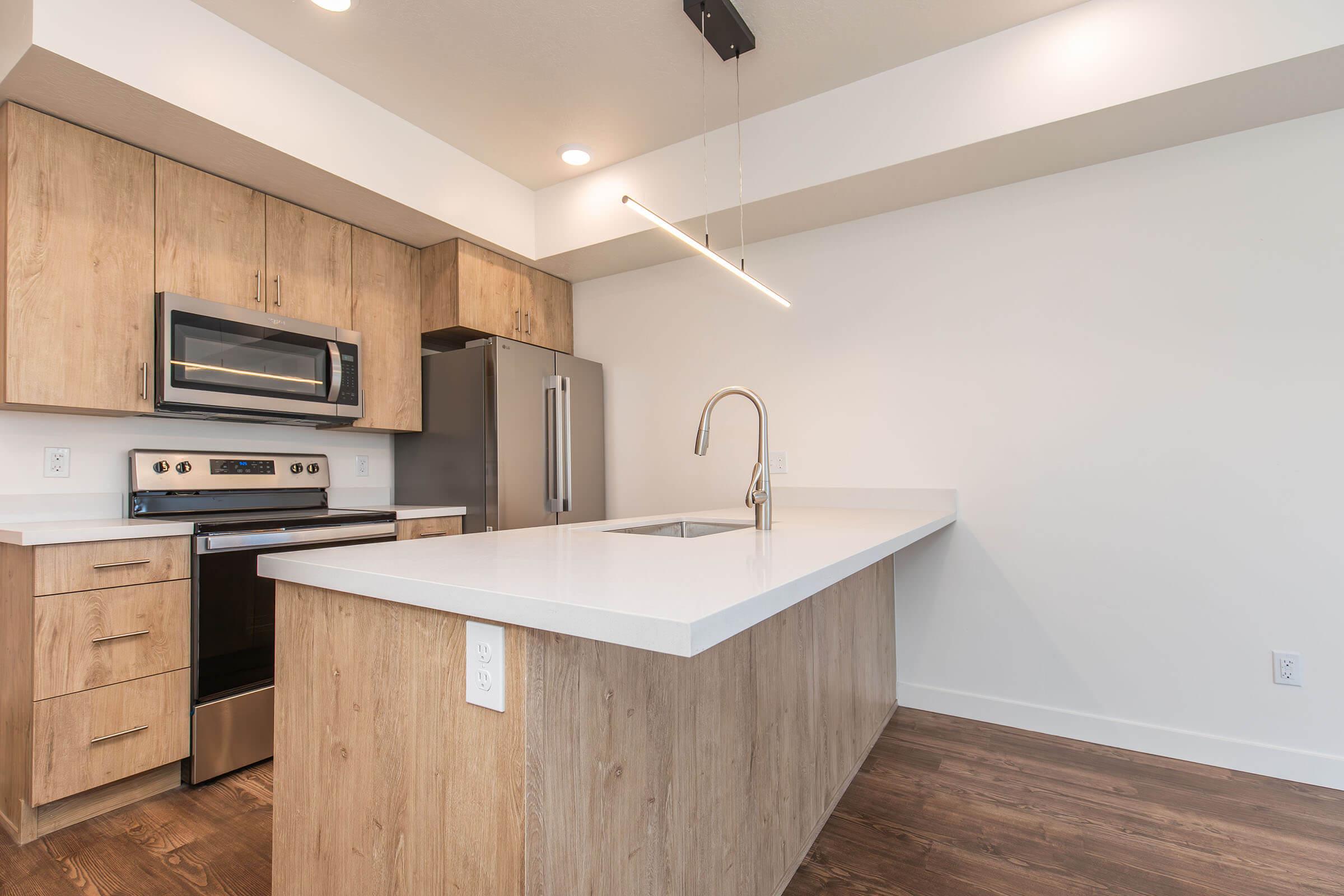 Modern kitchen featuring wooden cabinetry, stainless steel appliances including a microwave and oven, and a large island with a sleek white countertop. The backsplash is minimalistic, and the space is well-lit with recessed lighting and a pendant light above the island. Hardwood flooring completes the clean, contemporary look.