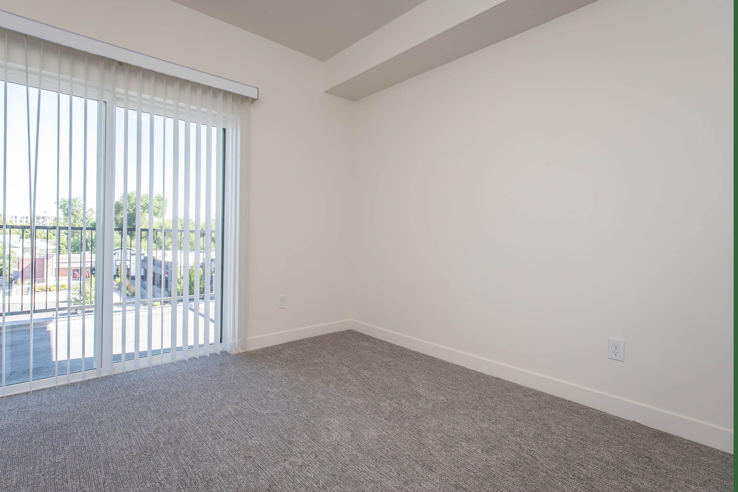 A spacious, empty room with light gray carpeting and white walls. It features a wall of vertical blinds that open to a balcony area. Natural light floods the space, creating a bright atmosphere. The corner of the room shows a small electrical outlet.