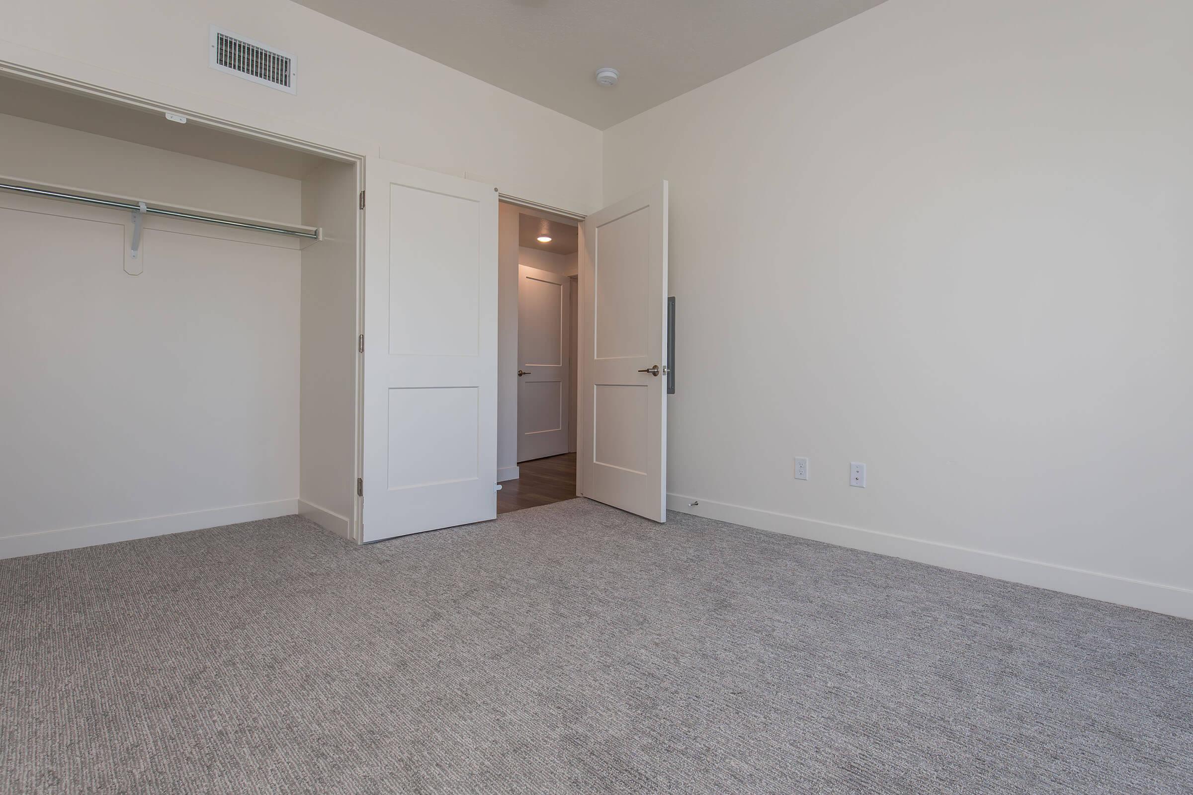 A spacious, empty room with light-colored walls and a plush carpet. There is a doorway leading to another area and a closet with a hanging rod on the left side. The overall atmosphere is bright and minimalistic, suitable for various purposes.