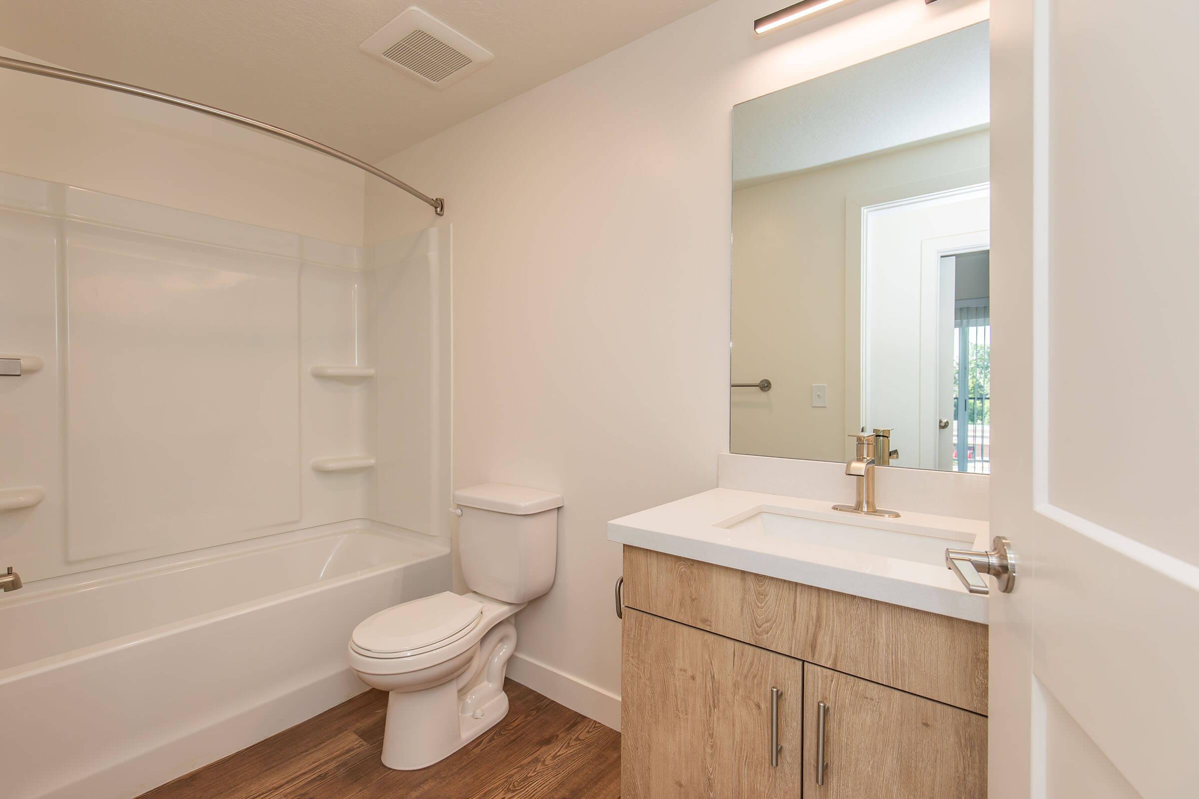 A modern bathroom featuring a bathtub with a shower curtain, a toilet, and a vanity with a sink. The walls are painted white, and there is a large mirror above the sink. The floor is made of wood-like material, and a portion of a doorway leading to another room is visible.