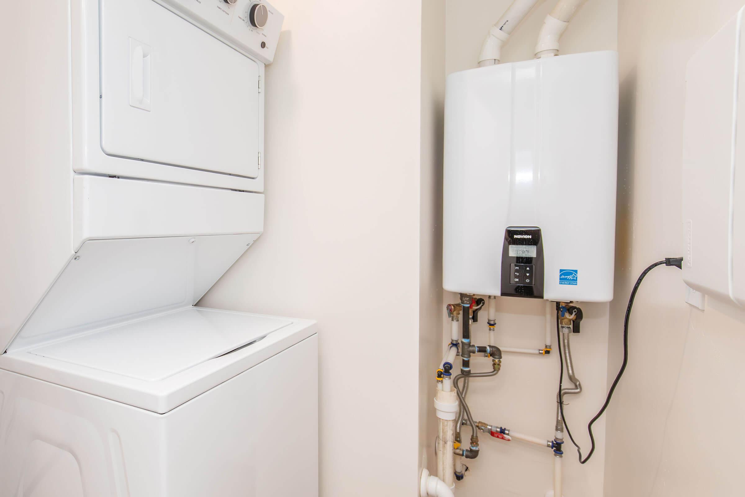 A laundry area featuring a stacked washer and dryer on the left and a white water heater mounted on the wall to the right, with visible plumbing connections and a clean, light-colored wall surrounding the appliances.