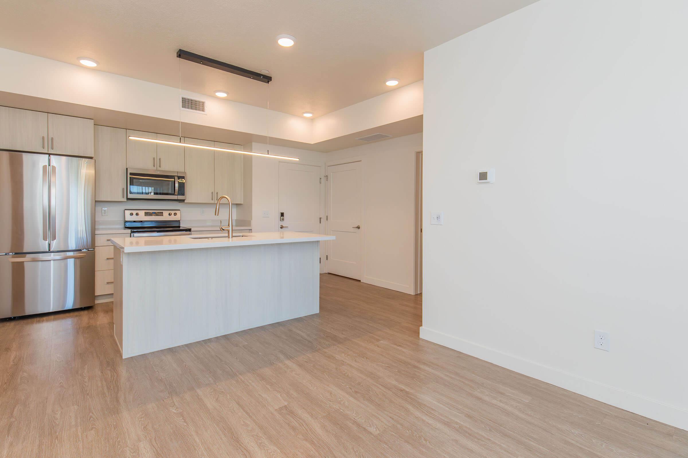 Modern kitchen with stainless steel appliances, including a fridge, oven, and microwave. An island with seating space is in the center, featuring a light countertop. The open layout connects to a bright living area with light-colored walls and wood-like flooring. Natural light enters through overhead fixtures.
