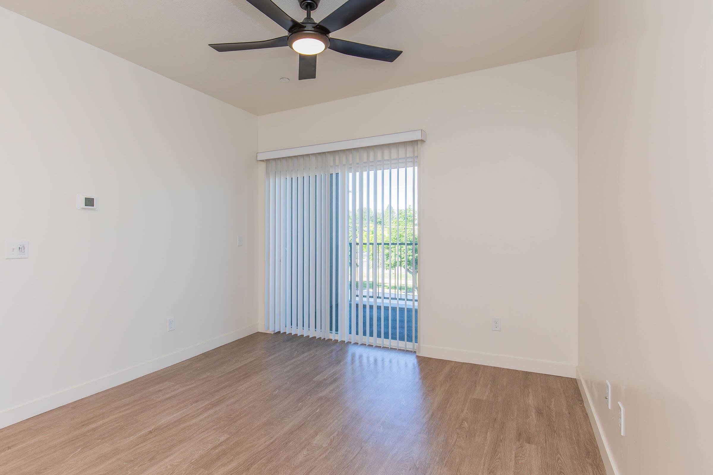A bright, empty room with light-colored walls and a ceiling fan. There are vertical blinds covering a sliding glass door that leads to a balcony. The floor is covered in light wood-like flooring, creating a clean and modern look.