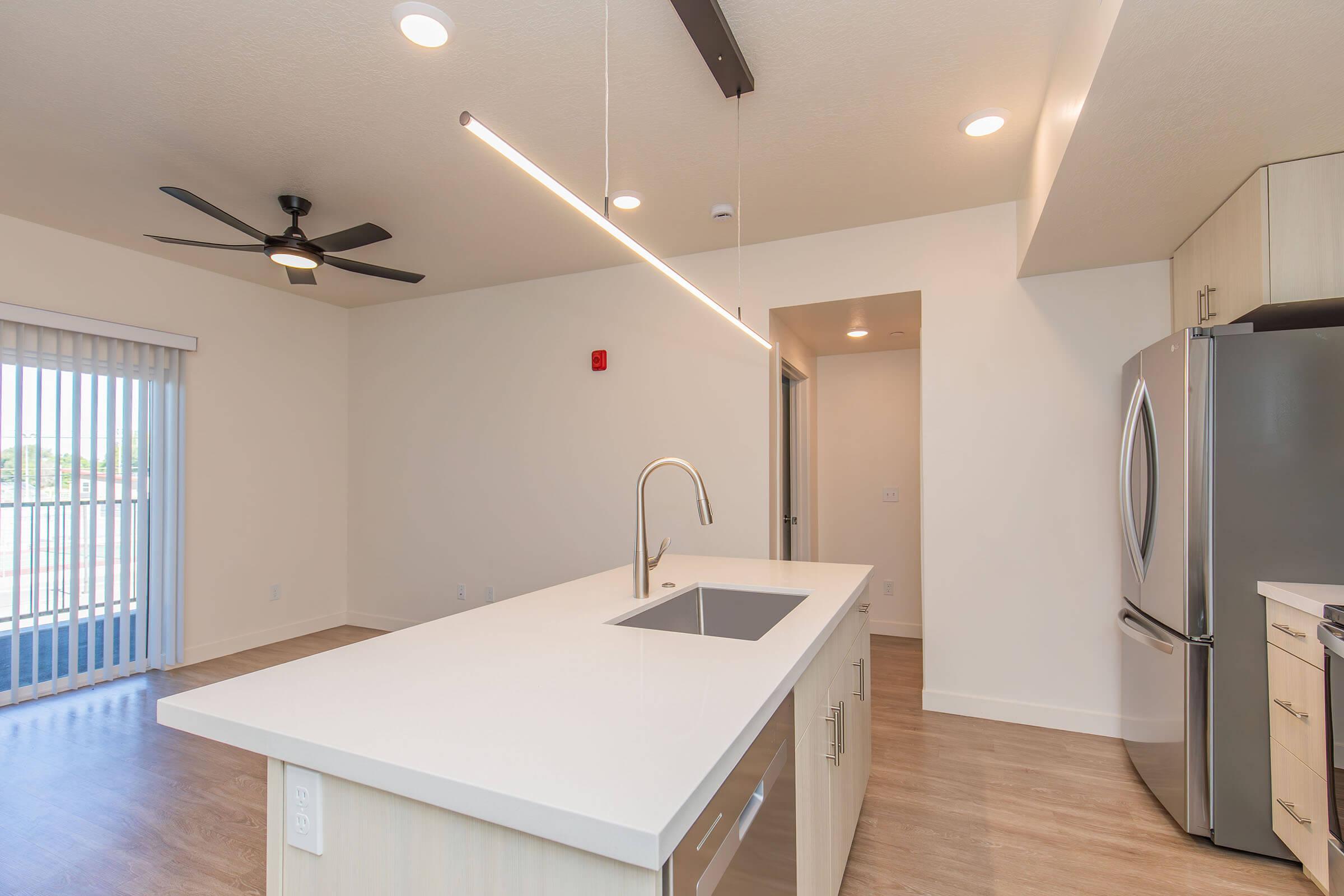 A modern kitchen featuring a large island with a sink, sleek white countertops, stainless steel appliances, and a ceiling fan. Large sliding glass doors provide natural light, and the space is complemented by light wood flooring and minimalist design elements.