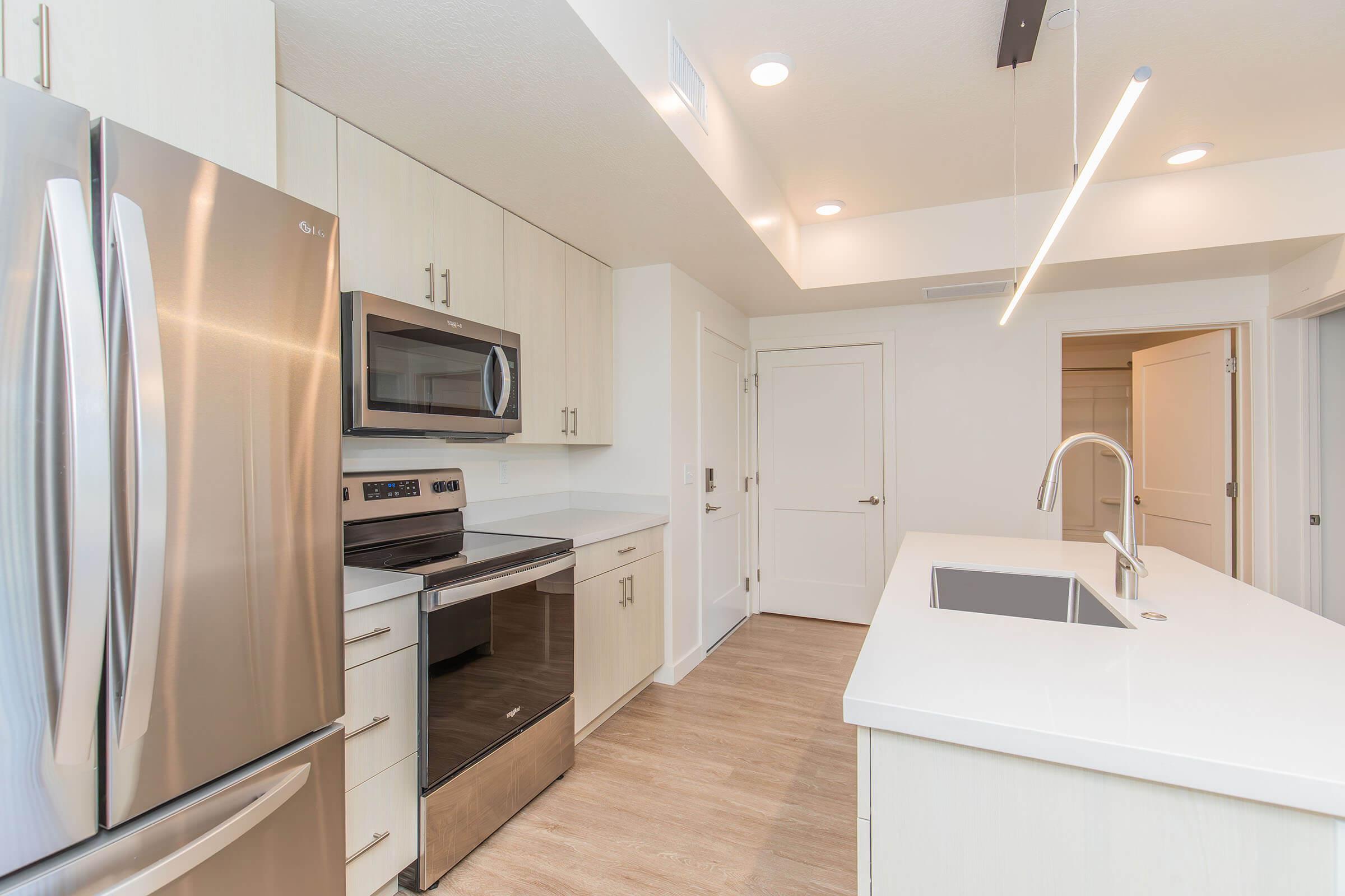 Modern kitchen featuring stainless steel appliances, including a refrigerator and oven. The cabinetry is light-colored, and the countertop is white with a modern sink. Soft lighting illuminates the space, and there is a minimalist design with an open layout. The room includes a door leading to another area.