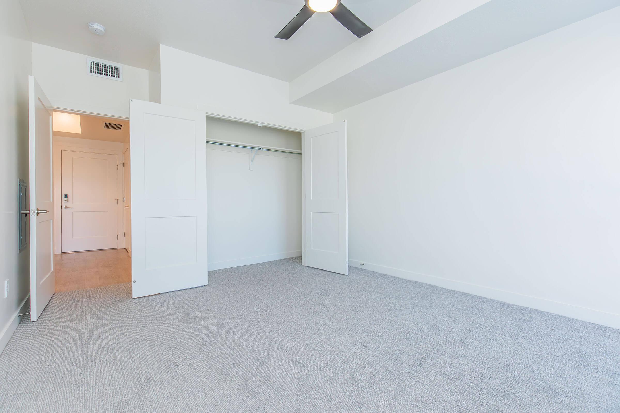 A spacious empty room with light-colored walls and a ceiling fan. The floor is covered with light carpet. A closet with open double doors is visible on one side, and an entrance door to another room is seen in the background. The overall atmosphere is bright and clean, suitable for various uses.