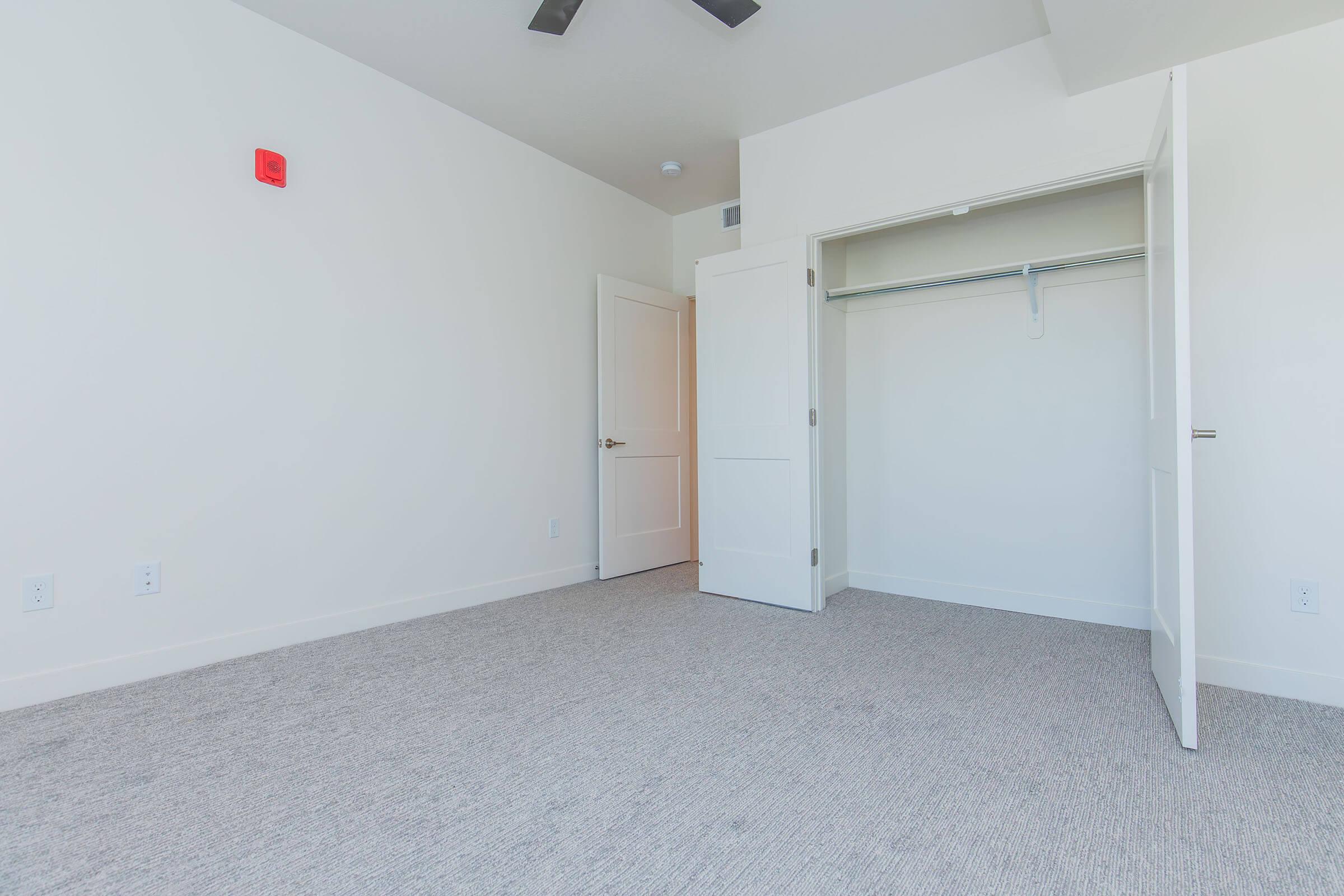 A bright, empty room featuring light-colored walls, a ceiling fan, and a carpeted floor. There is a door leading to another space and an open closet with a hanging rod. A red fire alarm is mounted on one wall, enhancing the room's safety features.