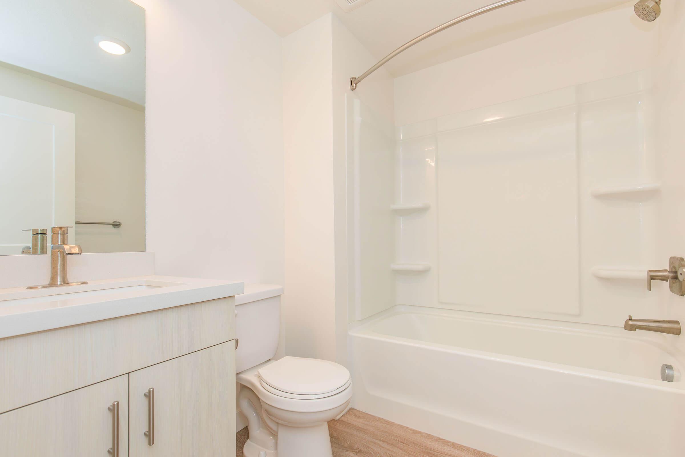 A clean and modern bathroom featuring a white bathtub with a shower curtain, a small white vanity with a sink, and a toilet. The walls are light-colored and the flooring is a light wood finish, creating a bright and spacious feel. Natural light enters from an unseen source.