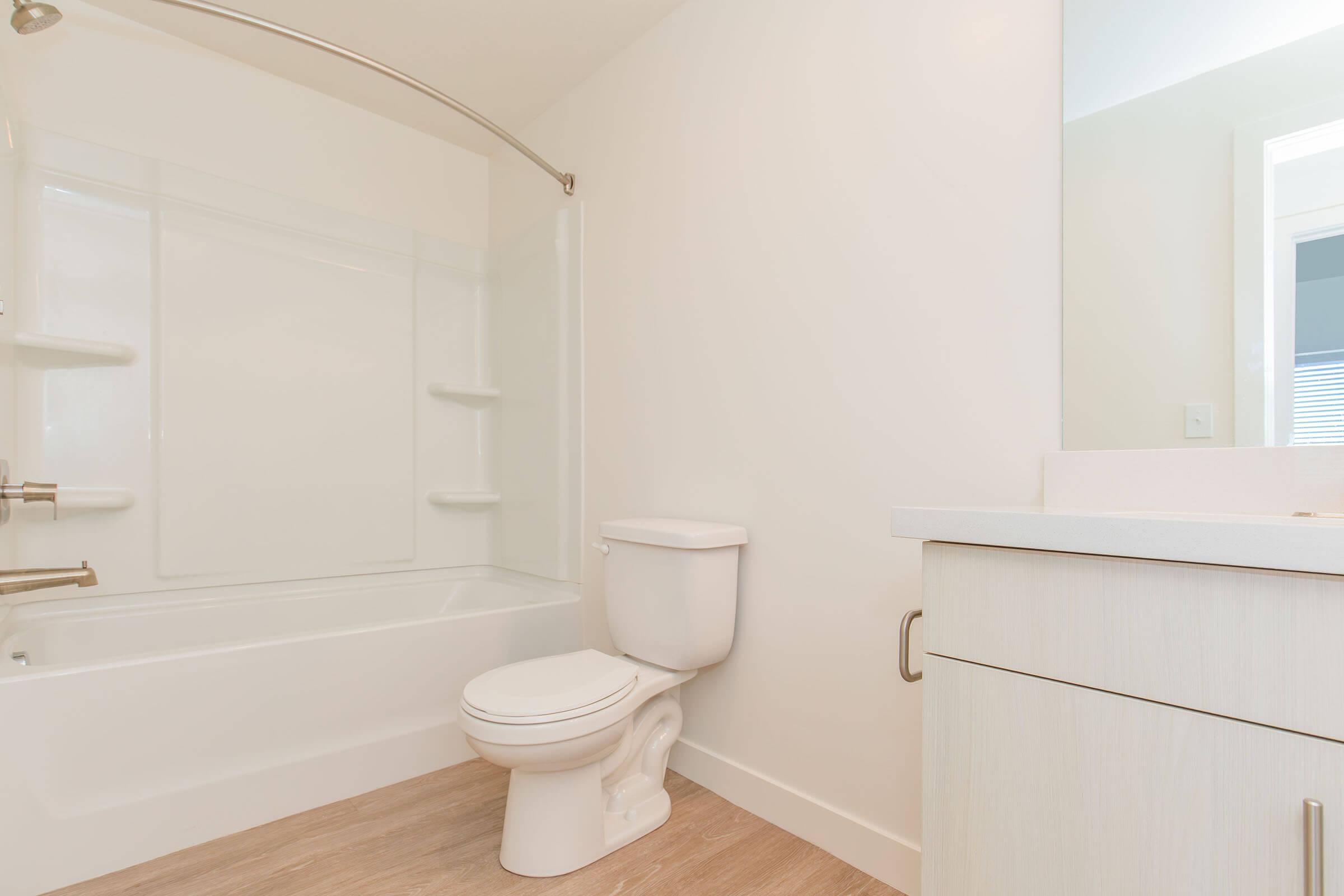 A clean, modern bathroom featuring a white bathtub with a shower, a toilet, and a vanity with a sink. The walls are light-colored, and the flooring is a light wood finish, creating a bright and spacious feel. Soft natural light illuminates the room from a nearby window.