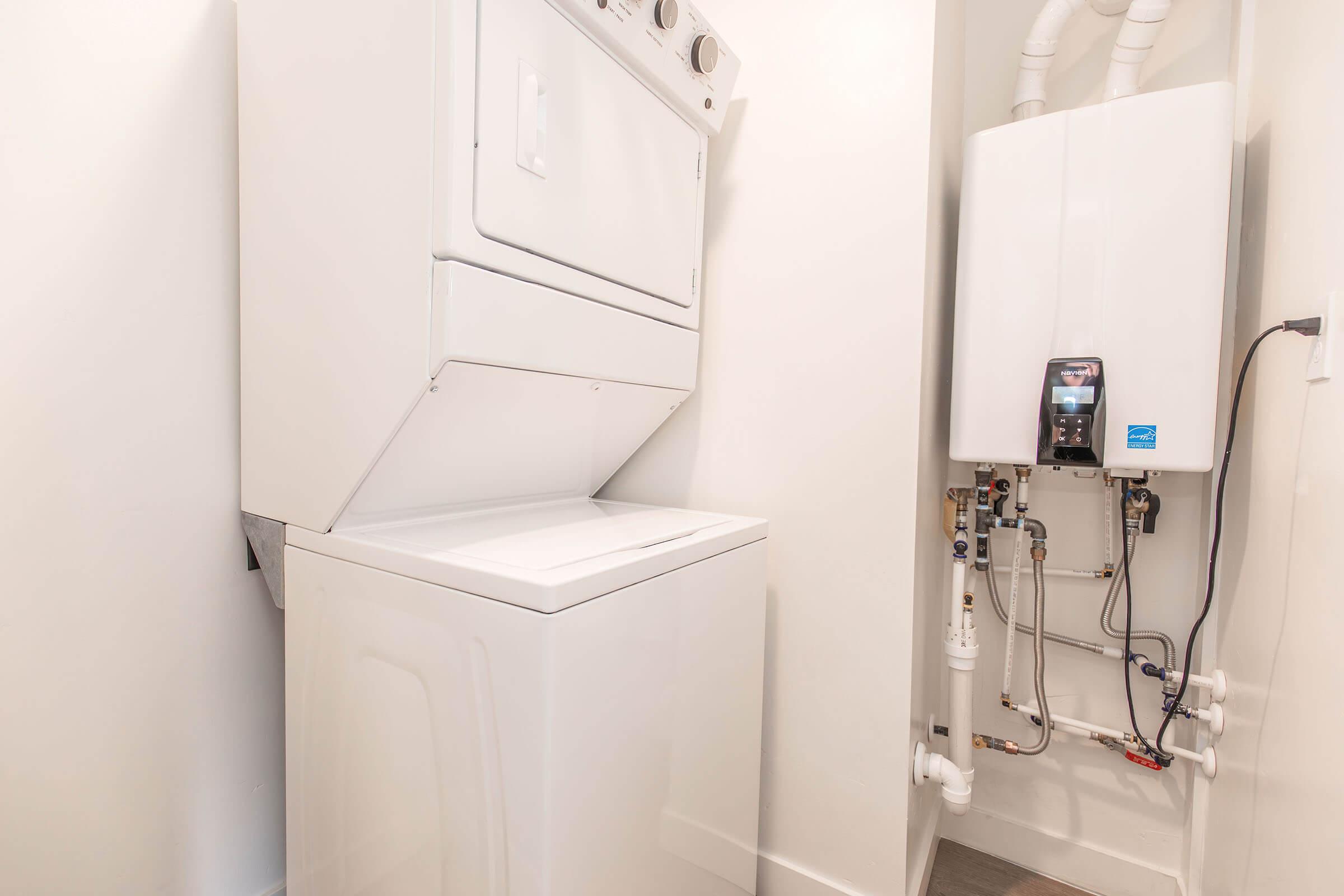 A compact laundry space featuring a stacked washer and dryer unit on the left and a tankless water heater mounted on the wall to the right, with visible plumbing connections and a clean, modern design. The walls are painted in a light color, enhancing the bright atmosphere of the room.