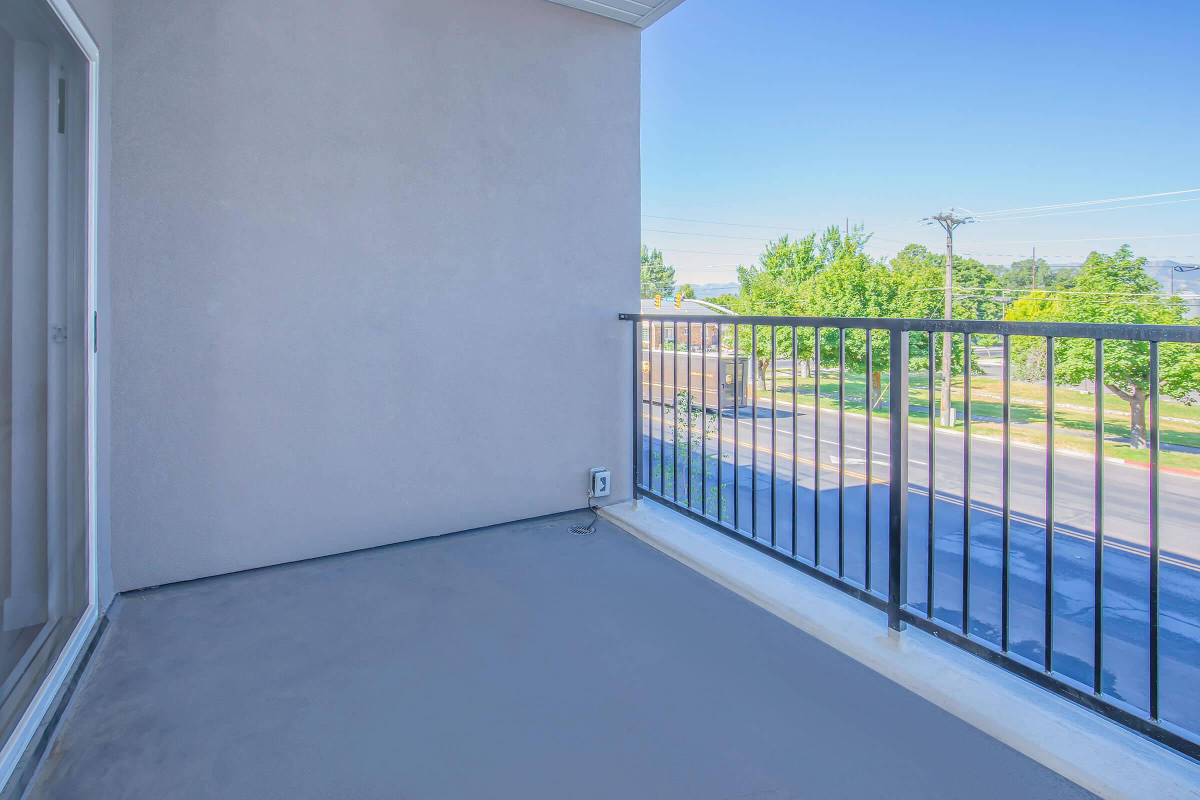 An empty balcony with a concrete floor and metal railing, overlooking a tree-lined street. The sky is clear and blue, with a few trees visible in the distance. The surroundings appear peaceful and inviting, suitable for relaxation or enjoying fresh air.