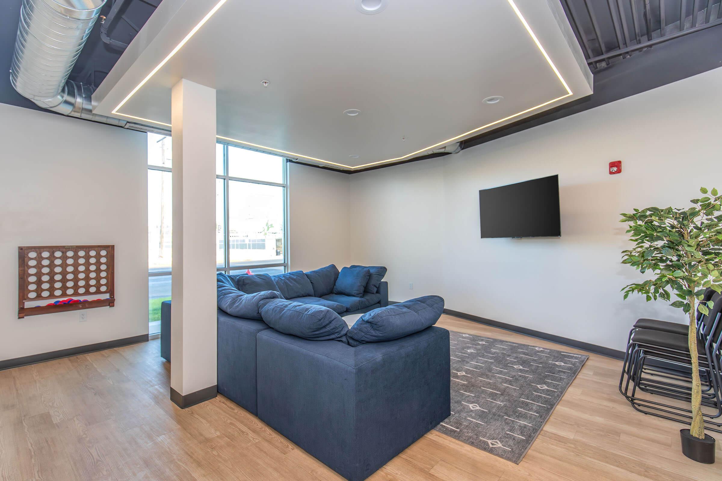 A modern living room featuring a large blue sectional sofa, a flat-screen TV mounted on the wall, and a decorative plant. The room has large windows allowing natural light, with a light wood floor and a patterned area rug, creating a cozy and inviting atmosphere.