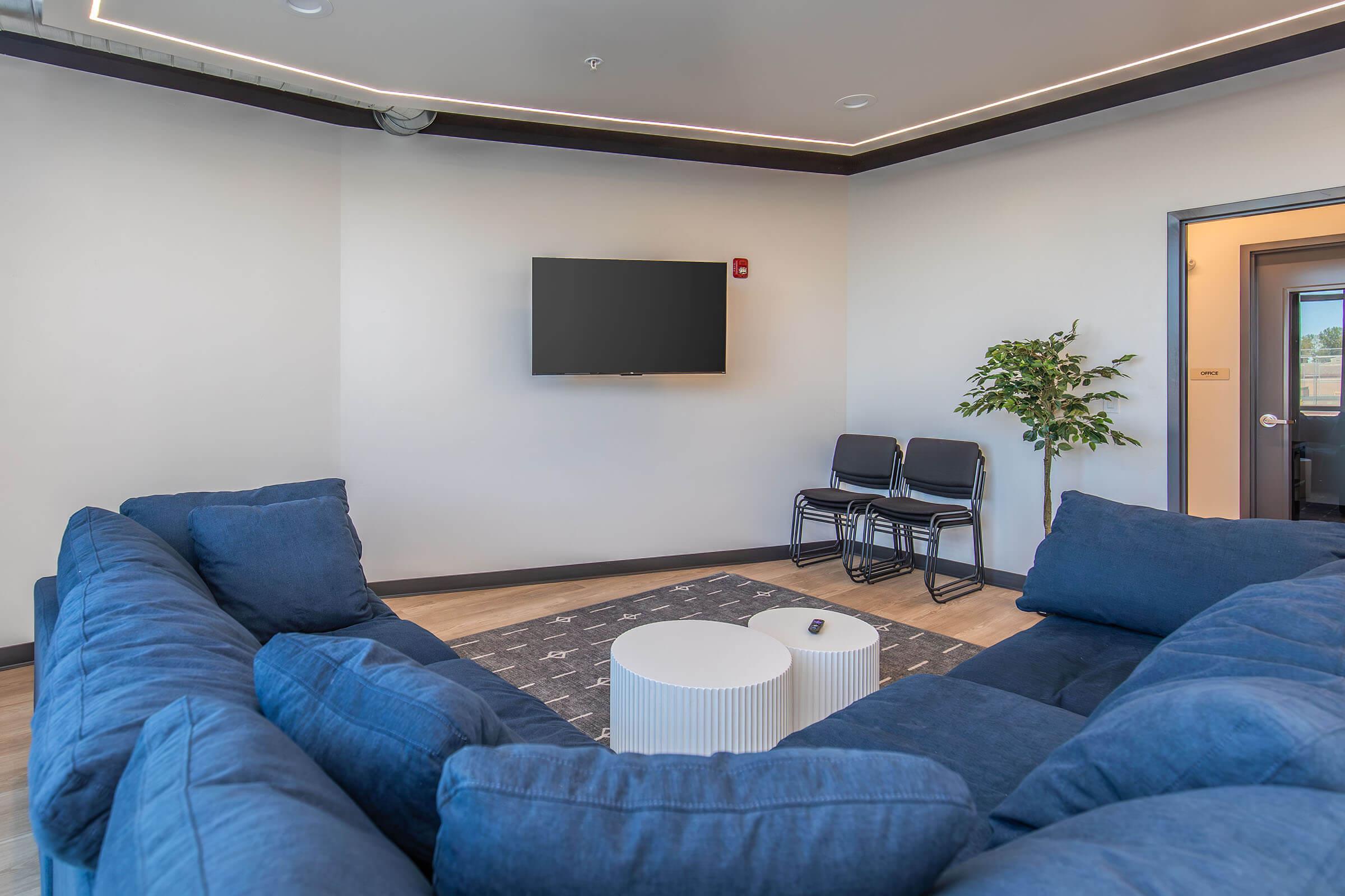 A modern lounge area featuring a large blue sectional sofa arranged in a circular layout, a round white coffee table in the center, a wall-mounted flat-screen TV, and two black chairs. A small green plant adds a touch of nature to the space, with neutral-colored walls and lighting that creates a welcoming atmosphere.