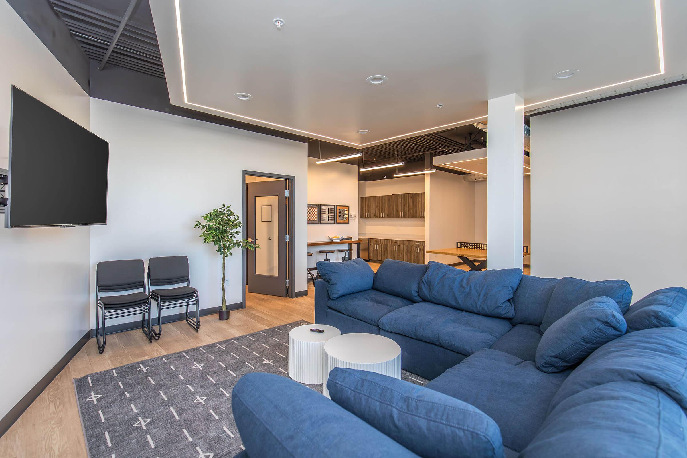 A modern living space featuring a blue sectional couch, a circular coffee table, and a patterned rug. There is a flat-screen TV mounted on the wall, a small potted plant, and chairs stacked in the corner. In the background, there is a doorway leading to another room with wooden cabinetry.