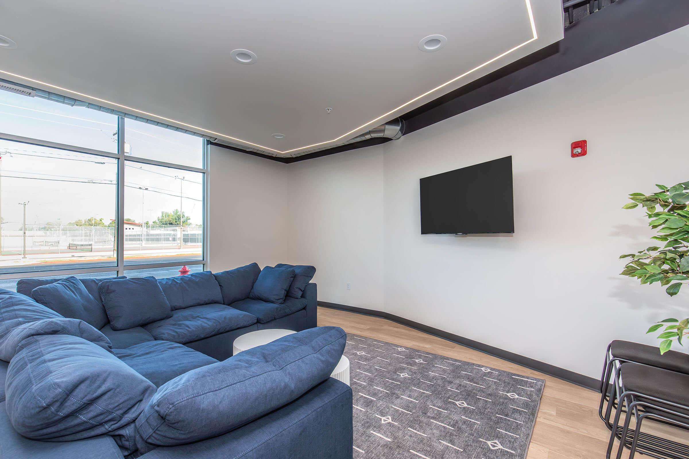 A modern living room featuring a large blue sectional sofa, a small round white coffee table, a mounted flat-screen TV on a light-colored wall, and a potted plant in the corner. The space has ample natural light from large windows, a sleek design, and minimalist decor.
