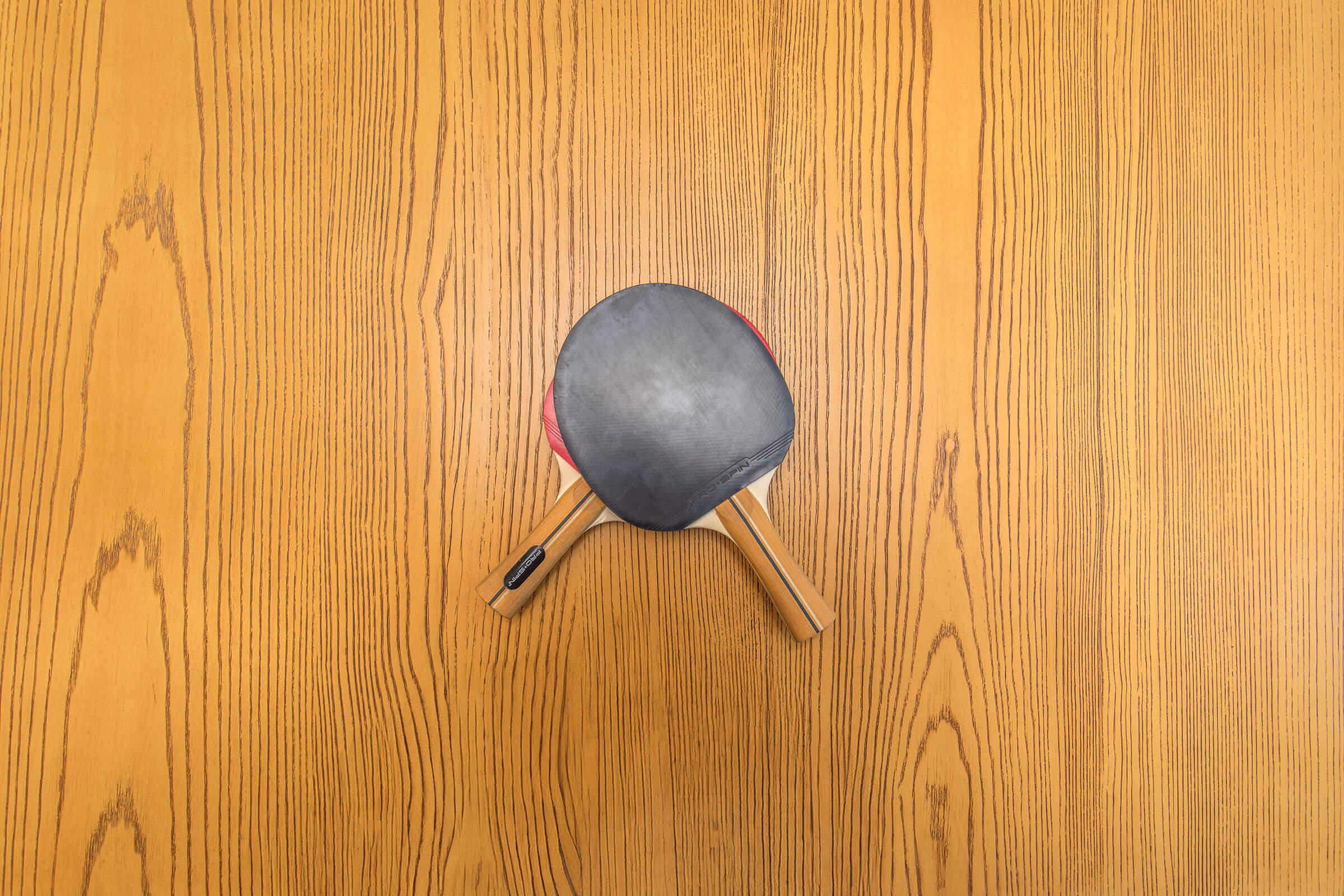 A top view of a single table tennis paddle resting on a wooden table surface. The paddle features a red rubber side and a black rubber side, set against the natural wood grain of the table.