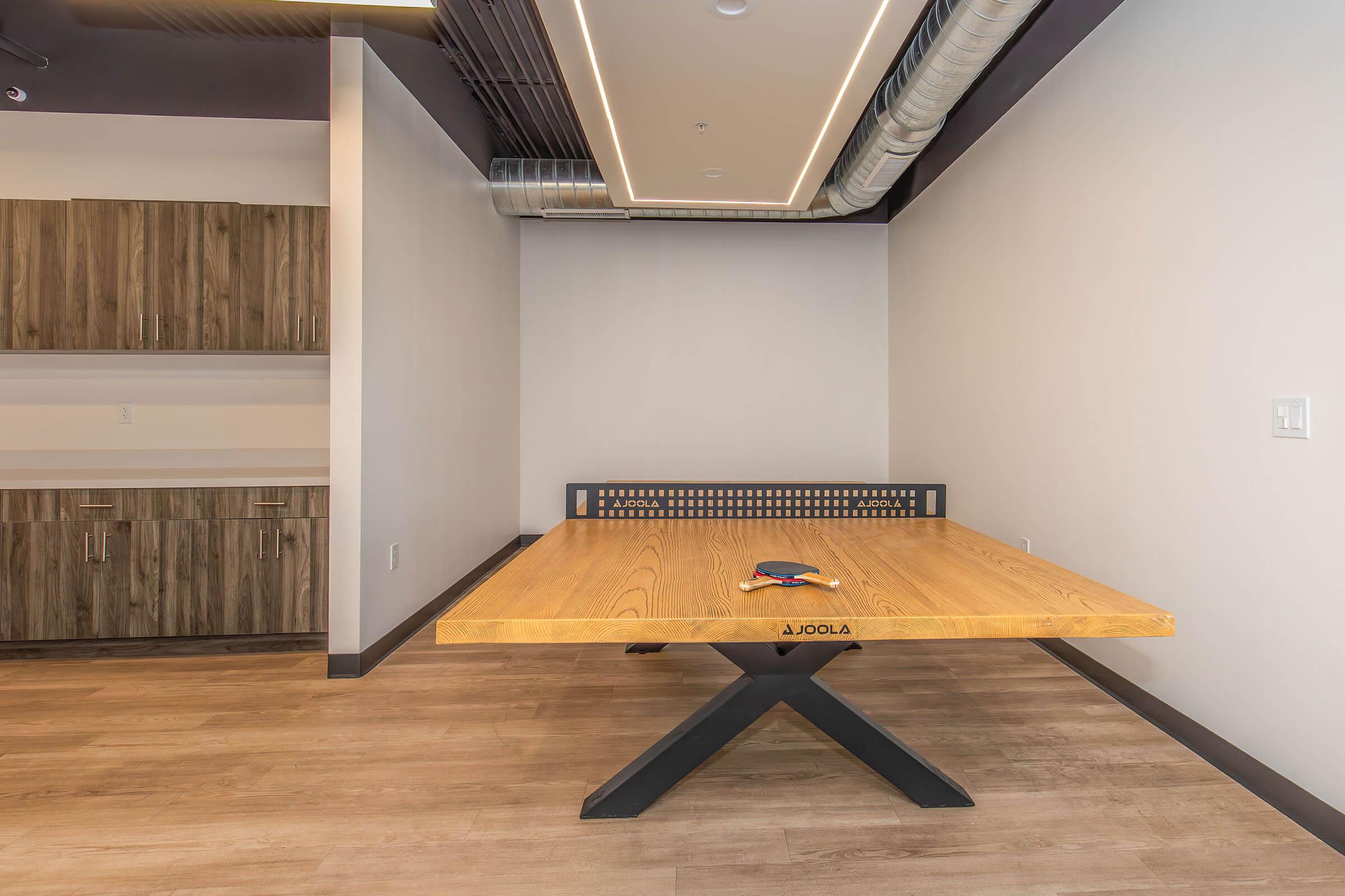 A modern room featuring a large wooden table with a sleek design, situated on a light wood floor. The walls are painted in neutral tones, and there are cabinets in the background. A ping pong paddle and ball are placed on the table, suggesting the space may be used for games or recreational activities.