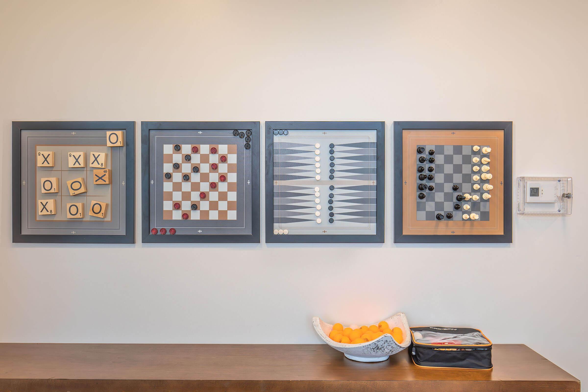 A wall display featuring four framed board games: Tic-Tac-Toe, Checkers, Backgammon, and Othello. Below the games, there is a bowl of oranges on a wooden surface alongside a small bag. The setting is bright and modern, with a simple, clean aesthetic.
